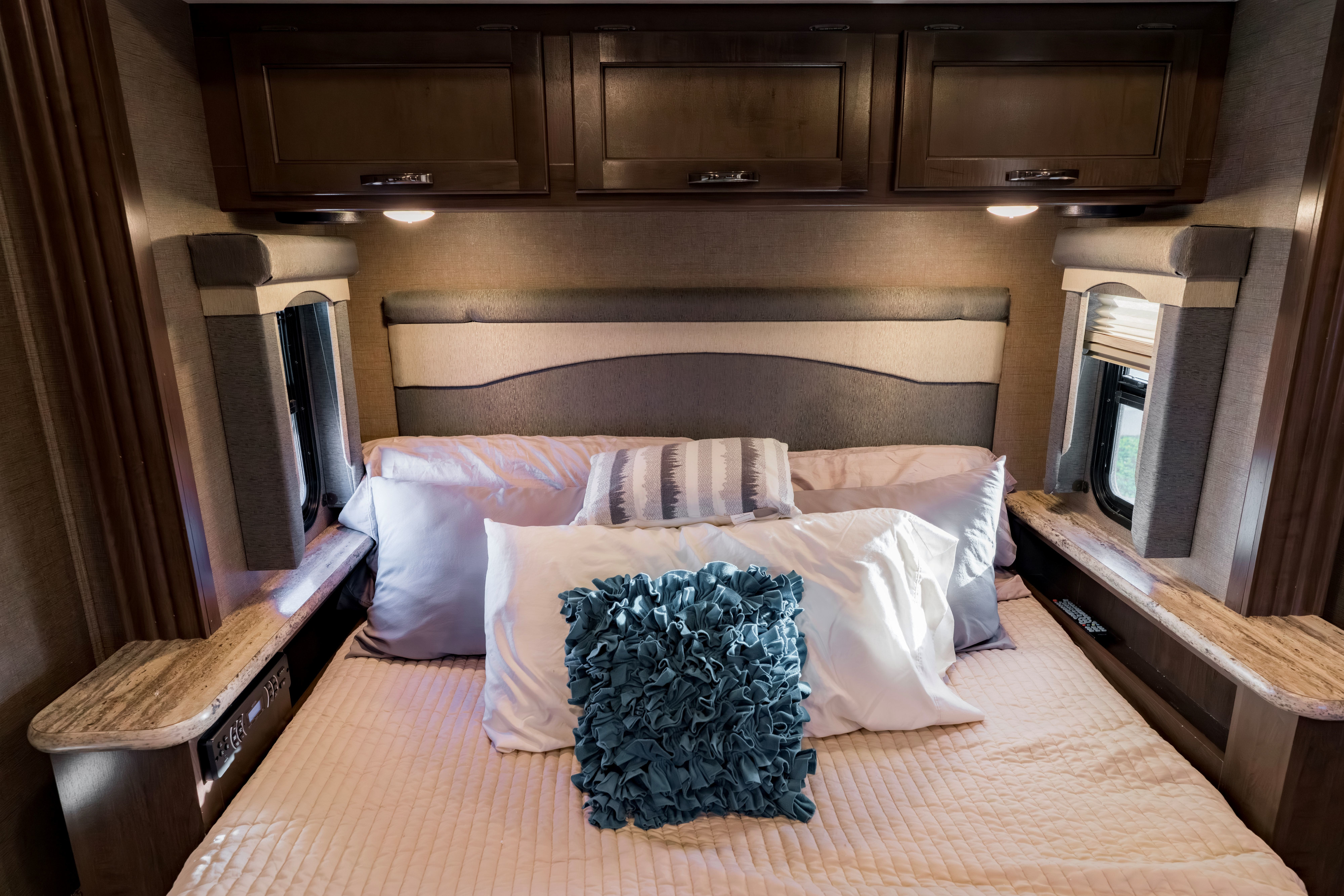 Cozy RV rental bedroom with a comfortable bed, textured headboard, overhead wooden cabinets, and built-in side shelves, softly lit.. Thor A.C.E. 2018