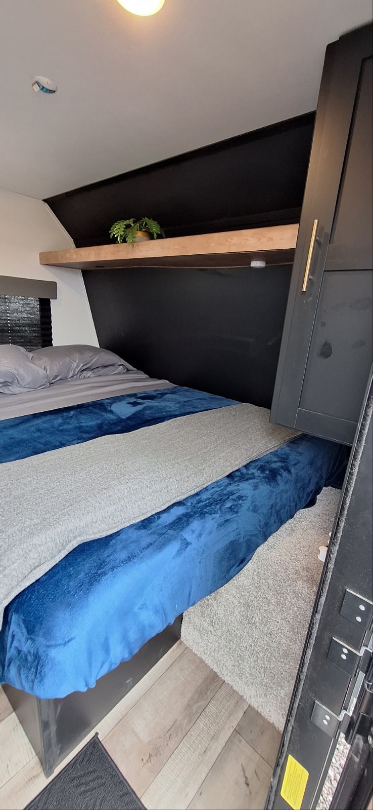 Inside a modern **rv rental** bedroom featuring a cozy bed with blue and grey blankets, a wood shelf, and dark cabinetry.. Springdale 260BH 2022