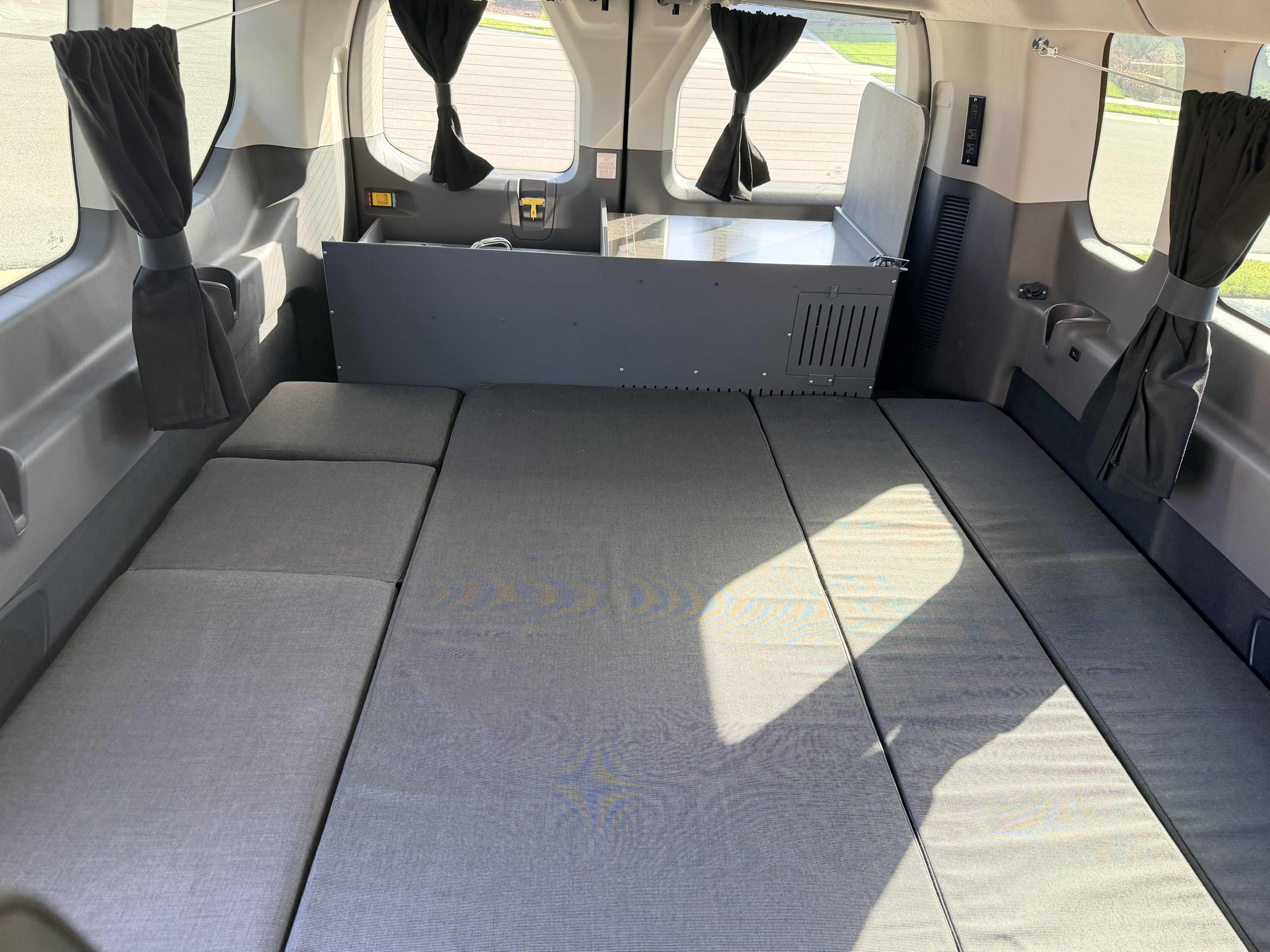 RV rental van interior featuring a grey modular bed, black curtains, and a compact kitchenette/storage unit.. Ford Transit 2017