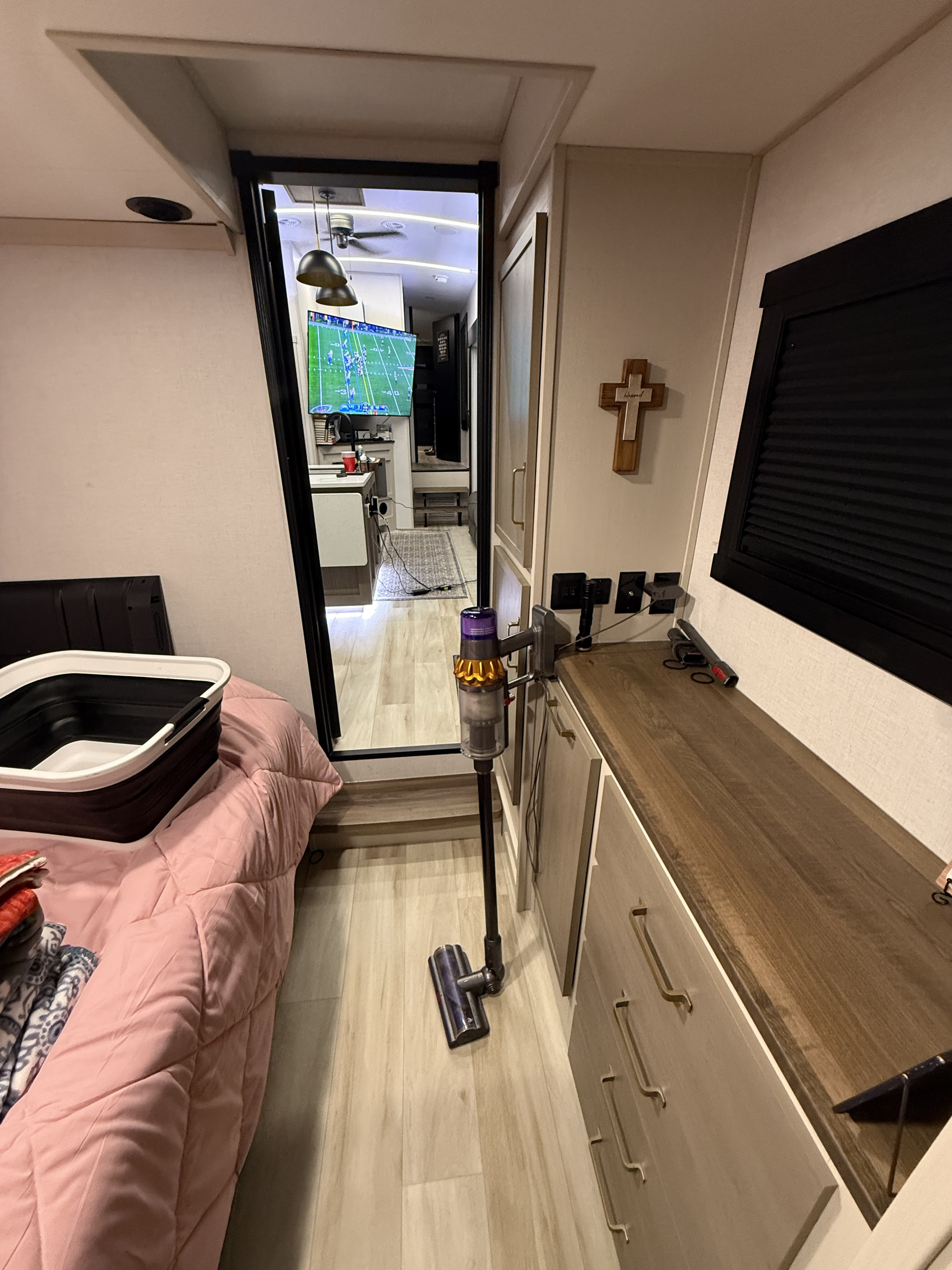 View inside an RV rental, showing a bedroom area with pink bedding, a Dyson vacuum, and a mirror reflecting a TV playing sports.. Forest River Salem Hemisphere 2025