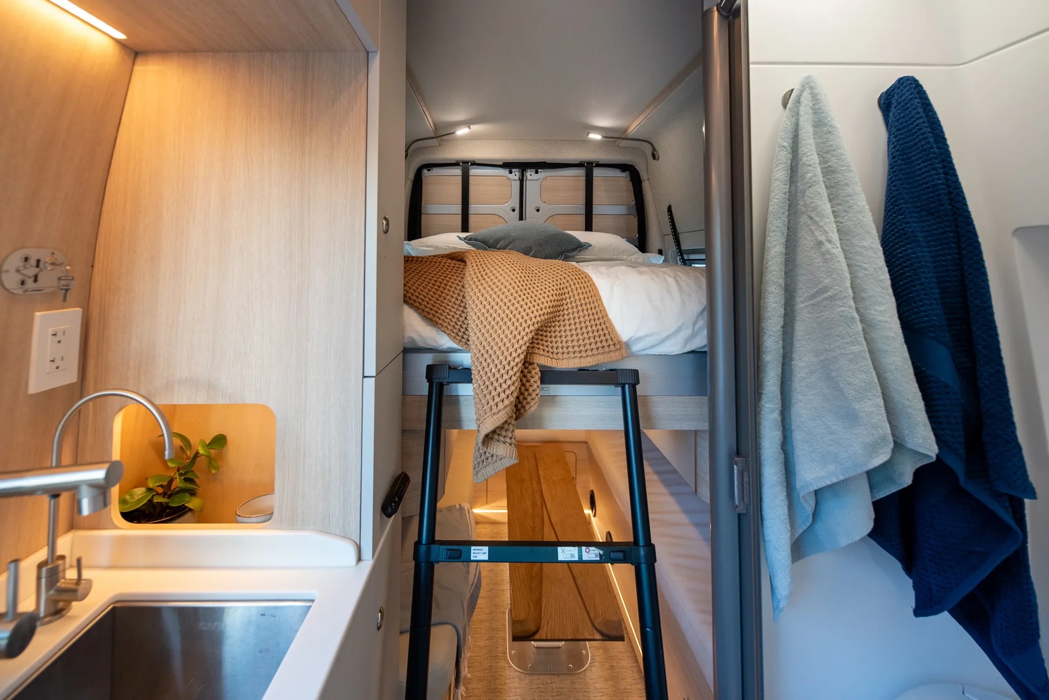 Functional RV rental interior: raised bed with ladder, compact kitchen sink, and cozy sleeping area. Towels on right.. Noovo Plus 2026