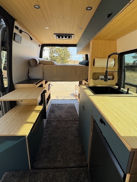Stylish RV rental interior: bed, modern kitchen with sink, light wood accents, & dark cabinetry along a plush rug runner.. Mercedes Sprinter 2026