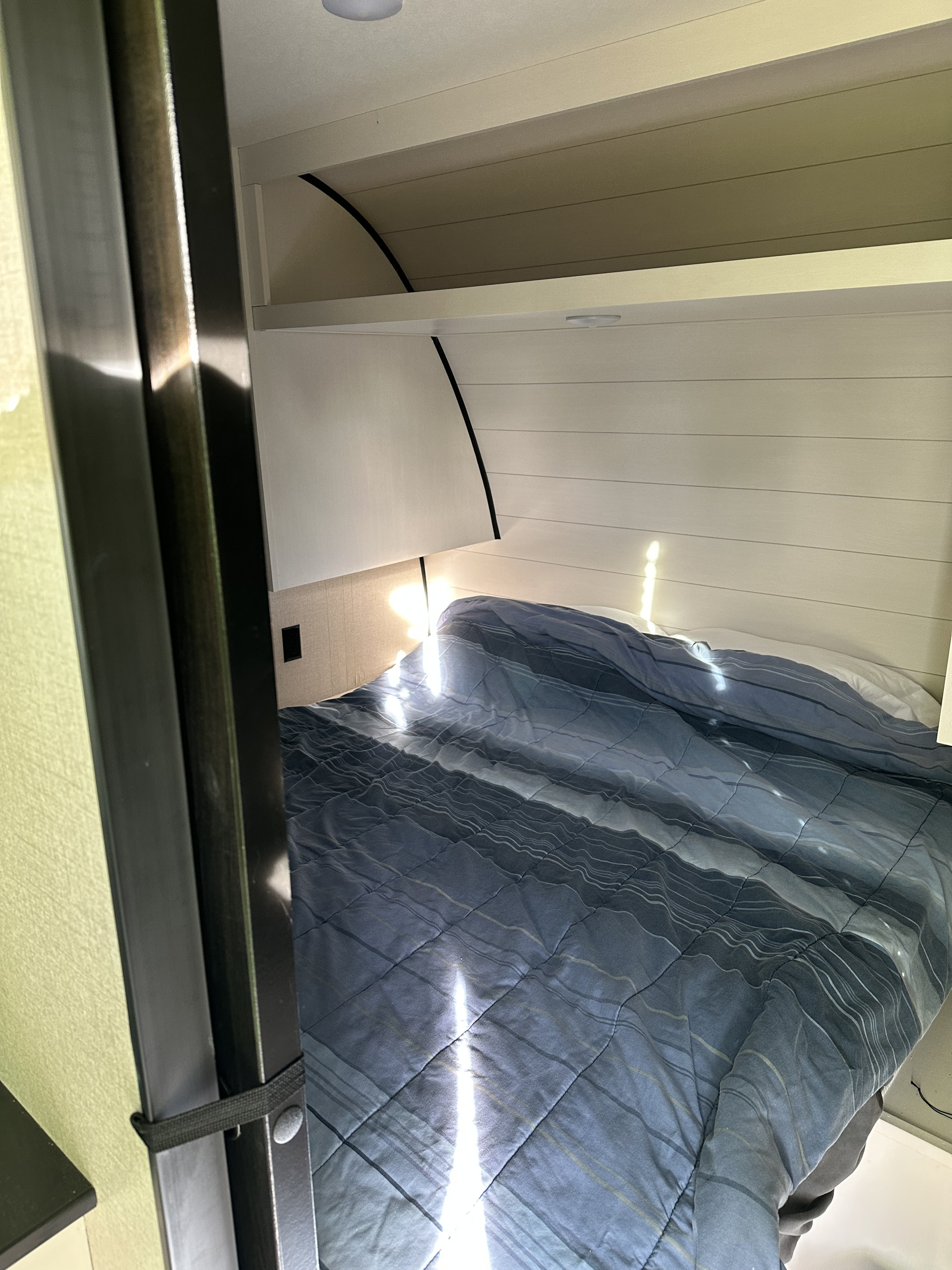 RV rental interior: a cozy bed with blue comforter, modern paneling, overhead shelf, and bright sunbeams.. Wayfinder RV GoPlay 26BHS 2024