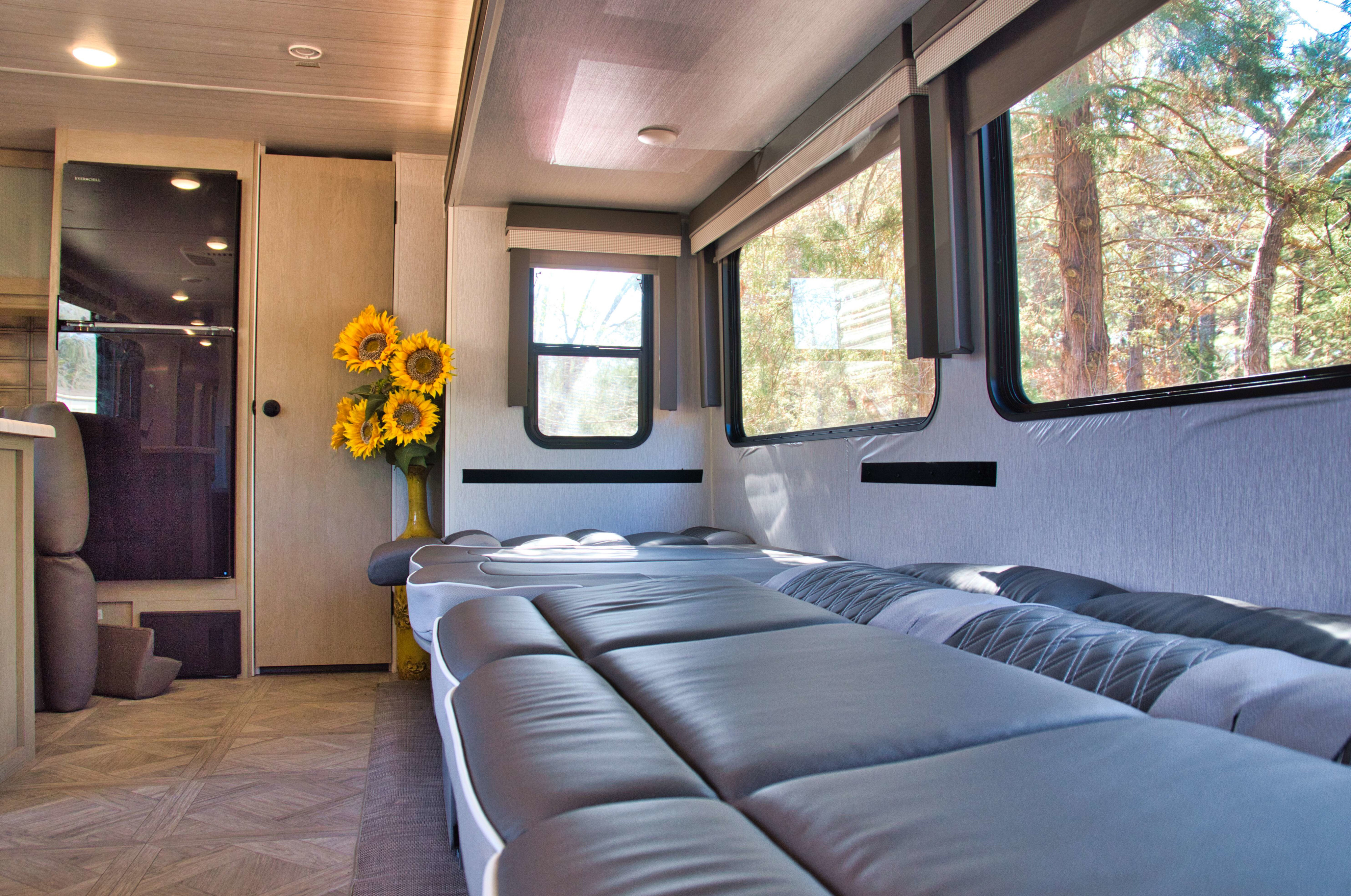 Cozy RV rental interior features a gray sofa bed, large windows with tree views, a refrigerator, and sunflowers in a vase.. Forest River Wildwood 2023
