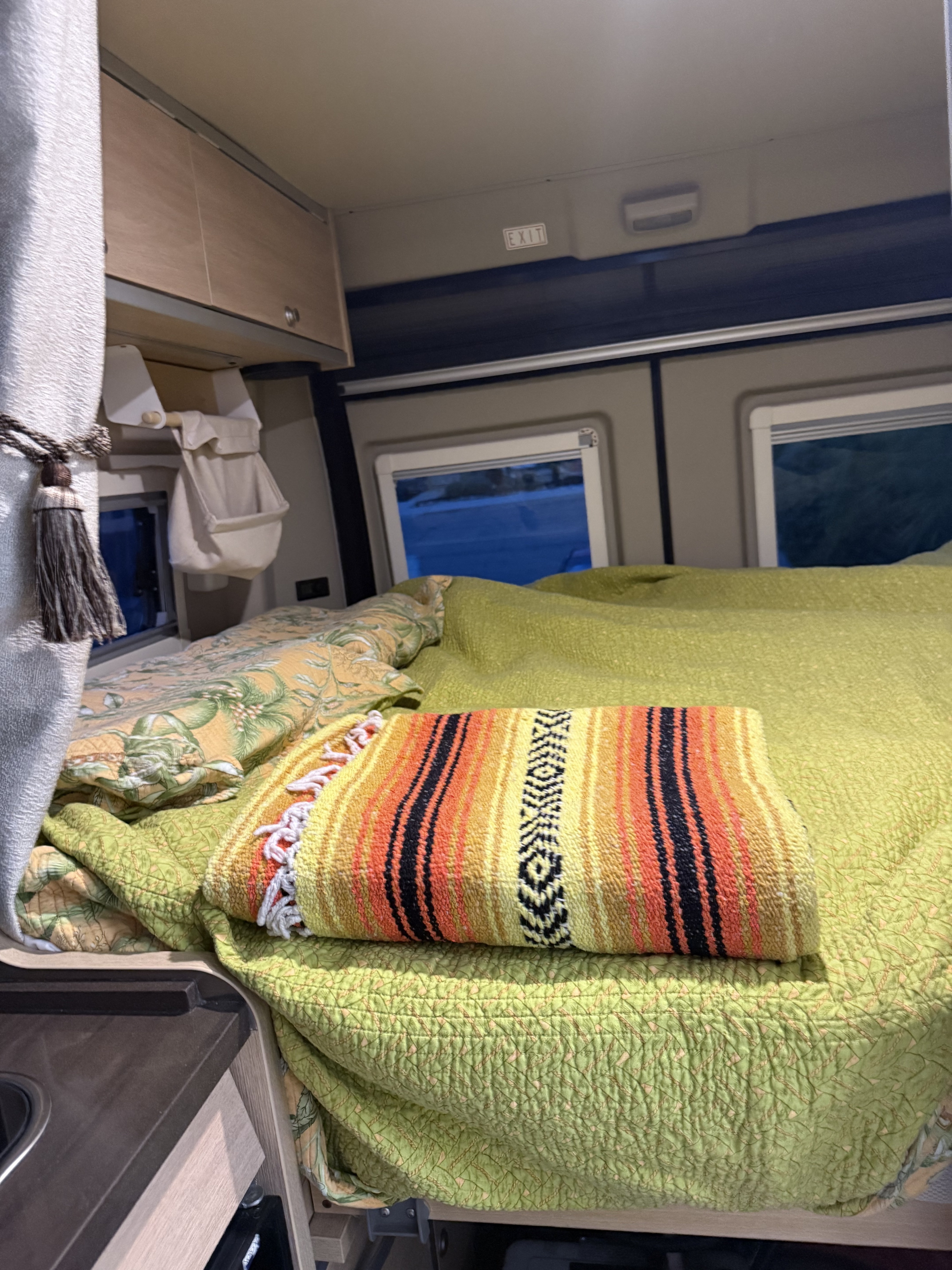 Inside a cozy **rv rental** camper van, featuring a vibrant bed with a green quilt and serape, overhead cabinets, and windows.. Hymer Hymer Class B 2019
