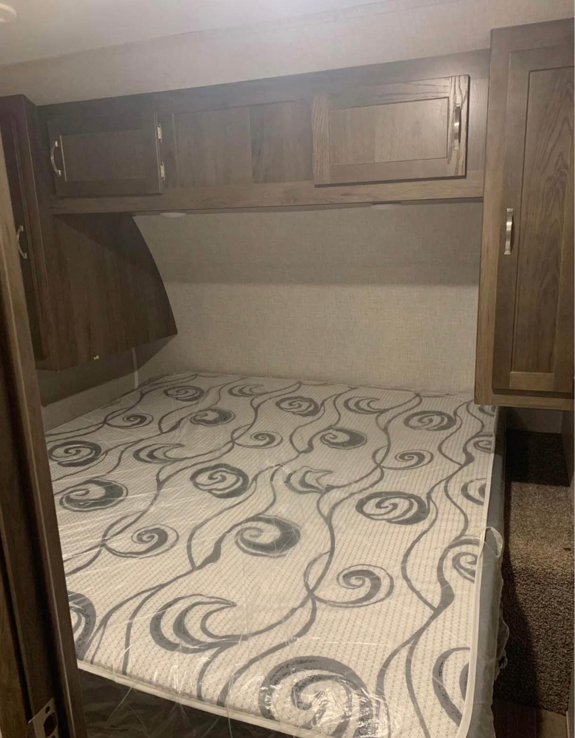 Experience comfort in this RV rental bedroom featuring a bed with a patterned mattress (still in plastic) and wooden overhead storage.. East to West Della Terra 2021