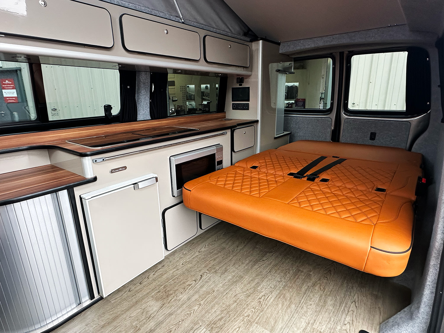 RV rental campervan interior: wood-finish kitchen with microwave, fridge, and an orange diamond-quilted bed.. Volkswagen Transporter 2024