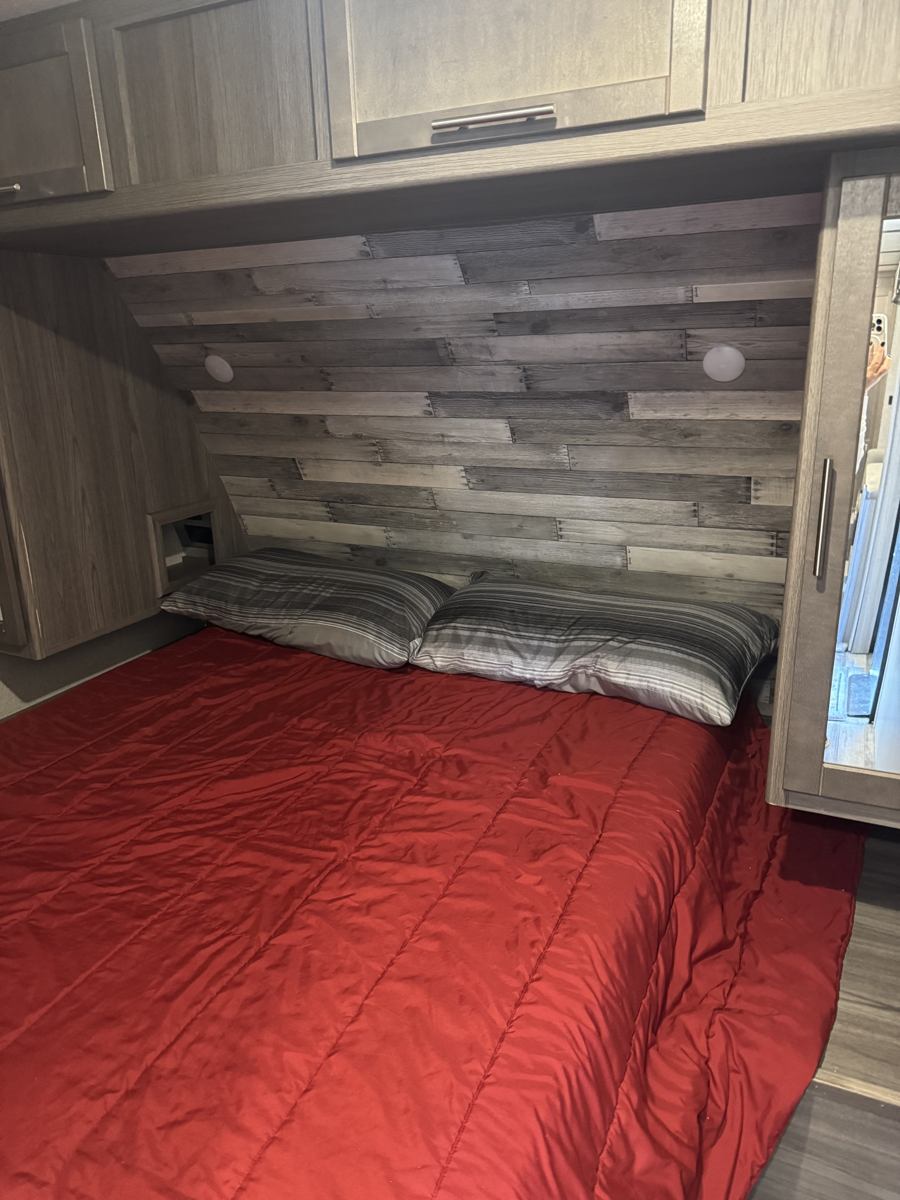 Cozy RV rental sleeping area featuring a red bed, rustic wood accent wall, and practical overhead cabinets.. Crossroads RV Volante 2021
