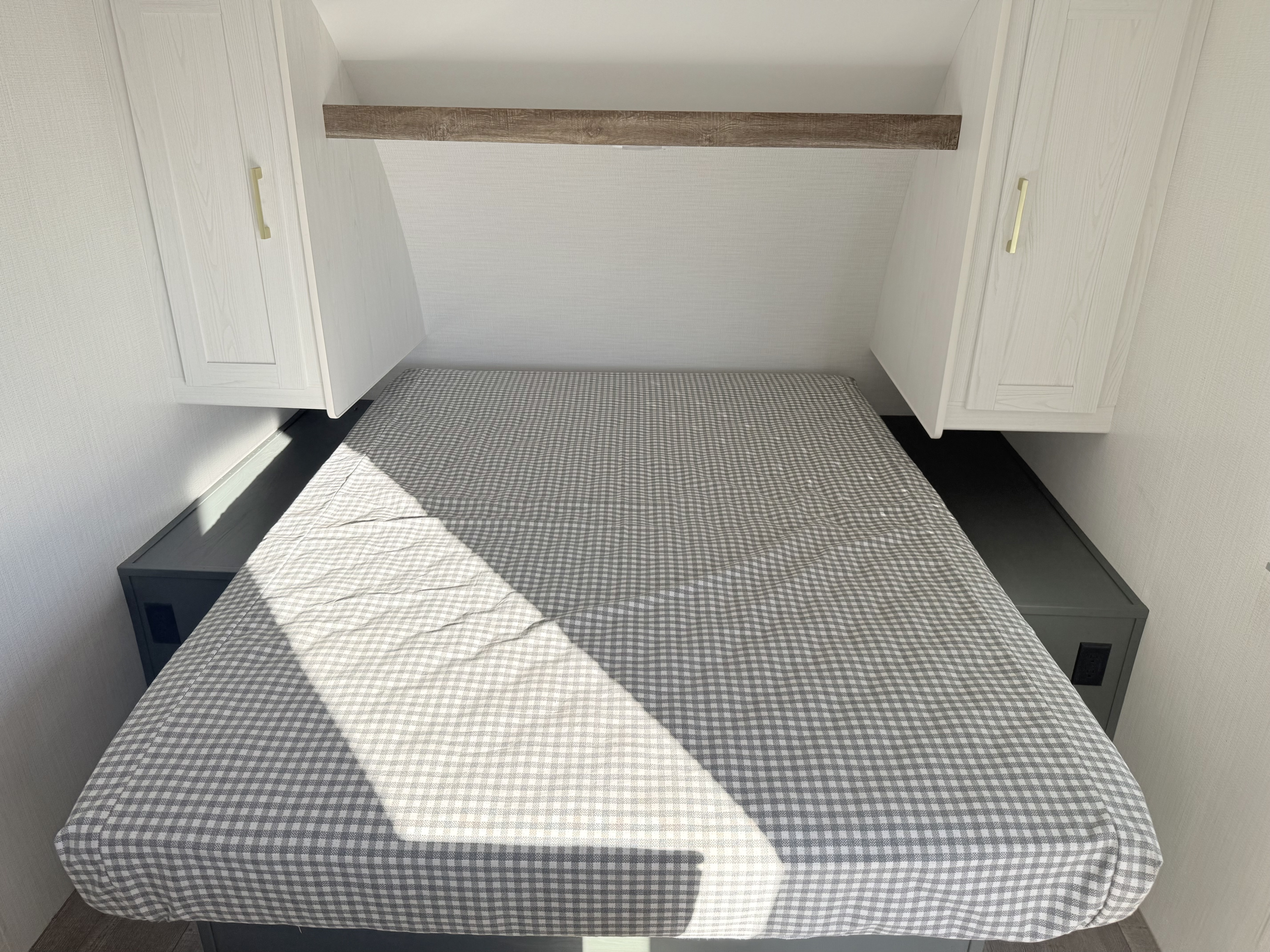 RV rental sleeping area: compact bedroom with gingham bed, light woodgrain cabinets, and storage. Sunlight adds warmth.. Heartland recreational vehicles 251BH-c 2024