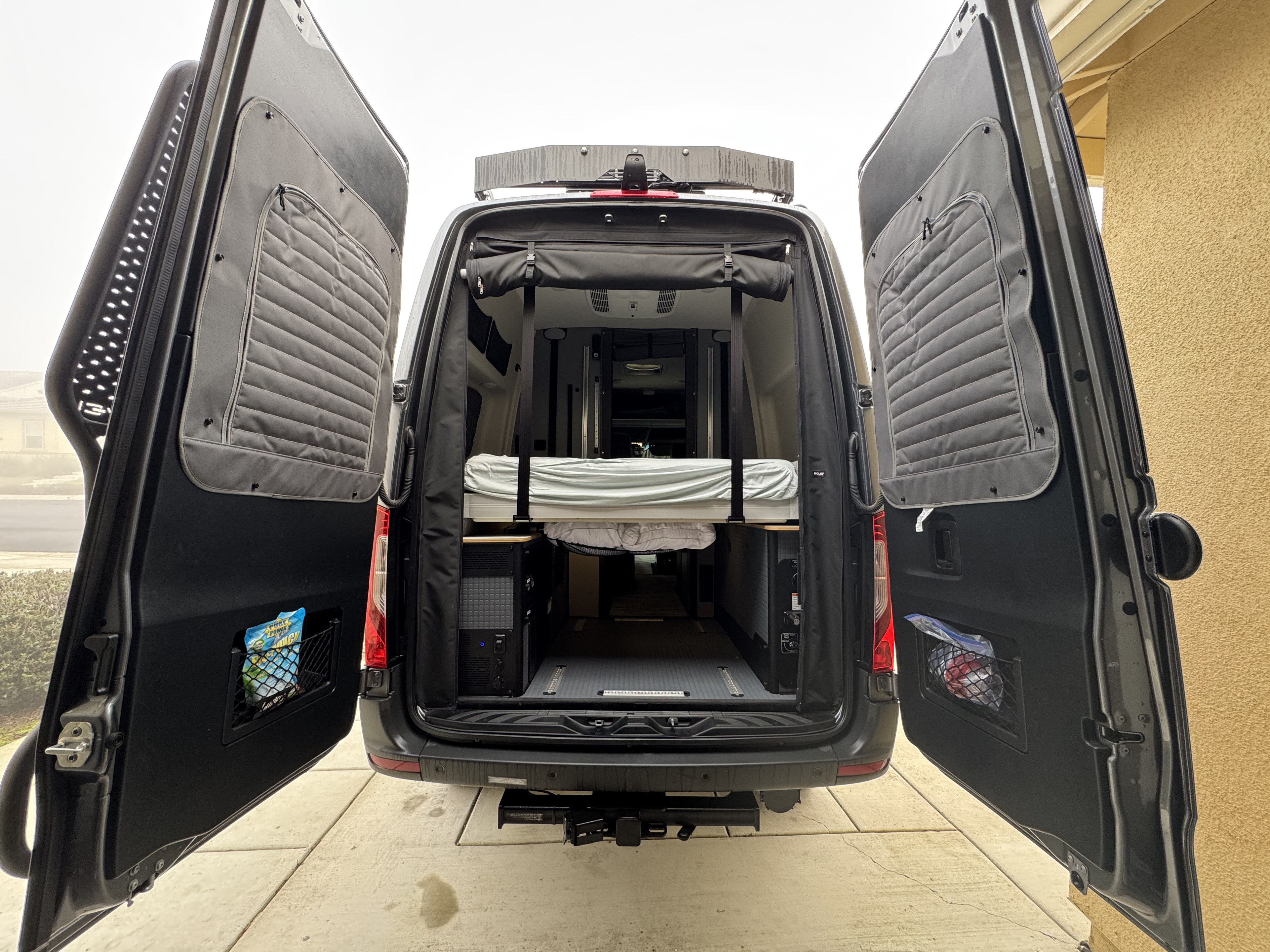 Rear view of an RV rental camper van with open doors, showcasing the interior bed, power system, and rear storage.. Winnebago Revel 2025