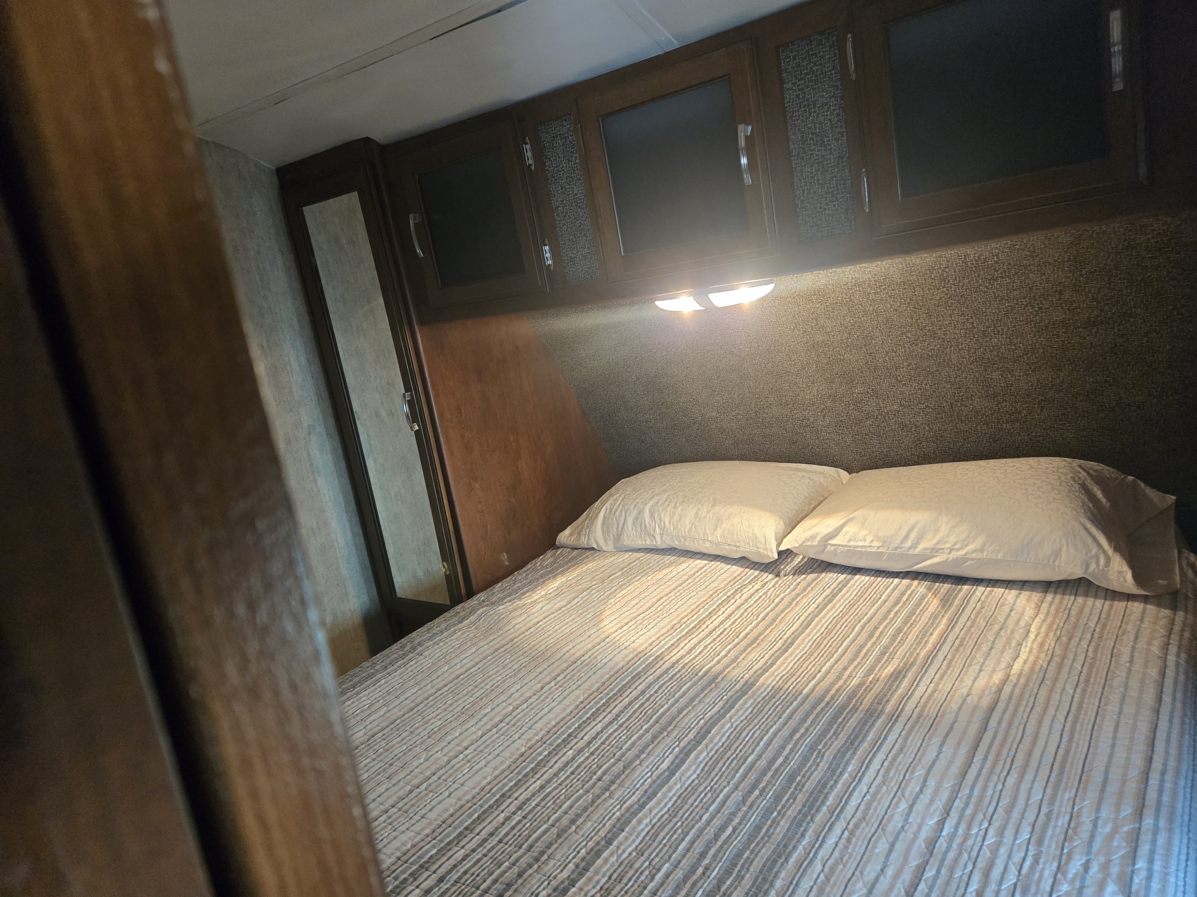 Inside an RV rental: a neat bedroom with a striped bed, white pillows, wood cabinets, and bright lights for comfort.. Keystone RV Passport SL 2020