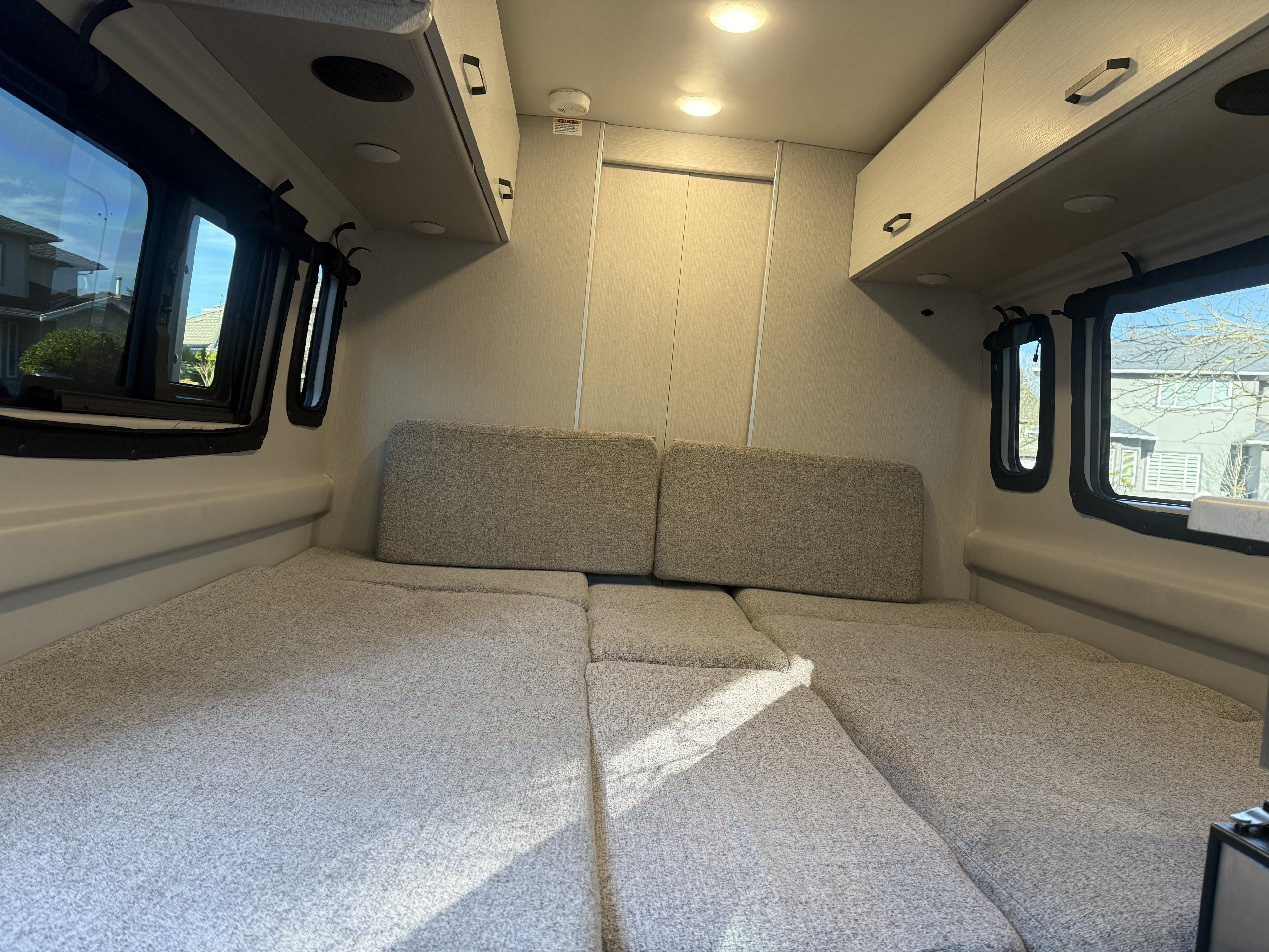 Bright rv rental interior: textured grey bed/lounge with windows & overhead cabinets, bathed in natural light.. Thor Motor Coach Ram promaster 2024