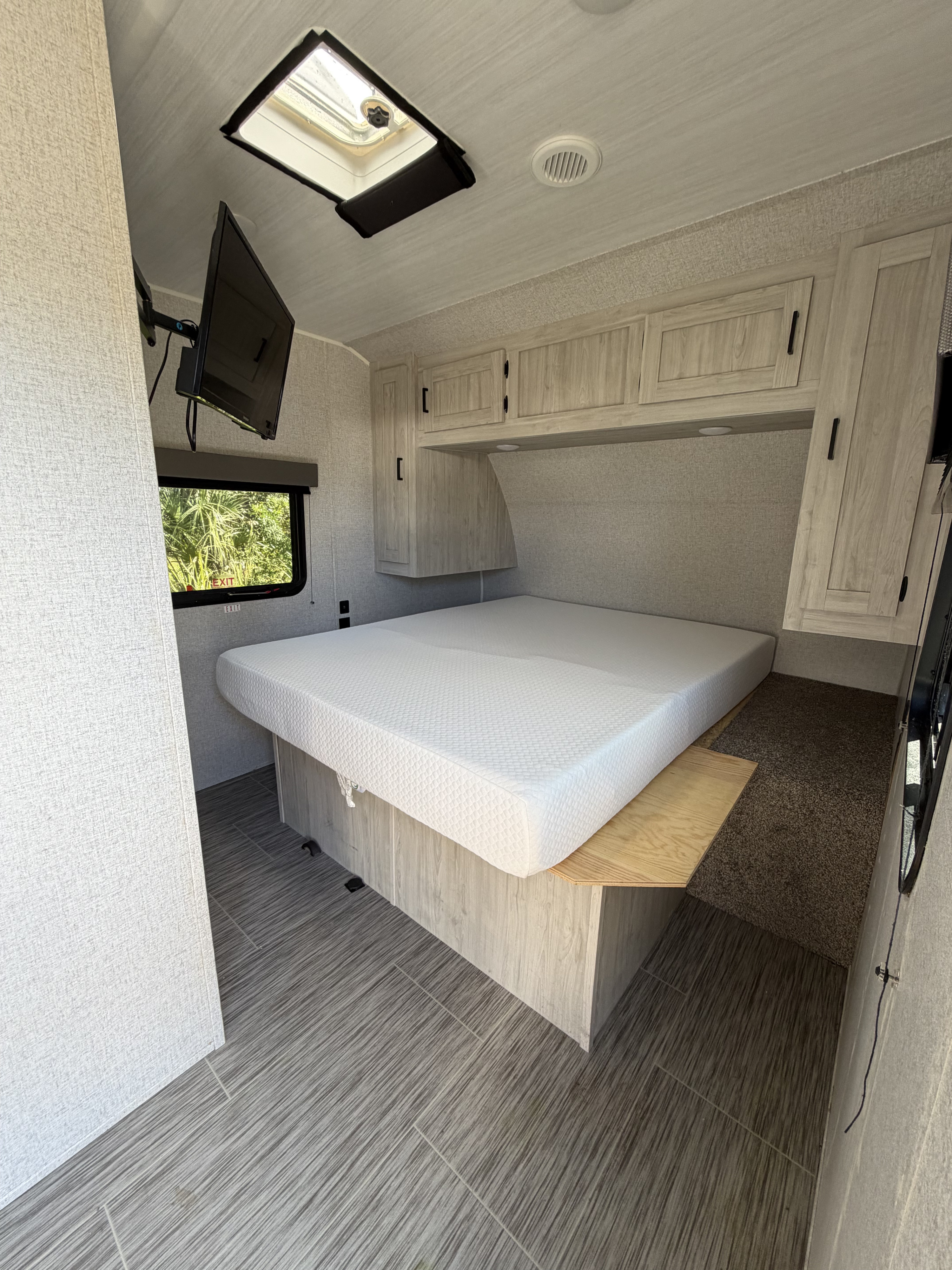 Modern RV rental bedroom with a comfy white mattress, light wood cabinetry, wall-mounted TV, window, and ceiling details.. East to West Silver Lake 2023