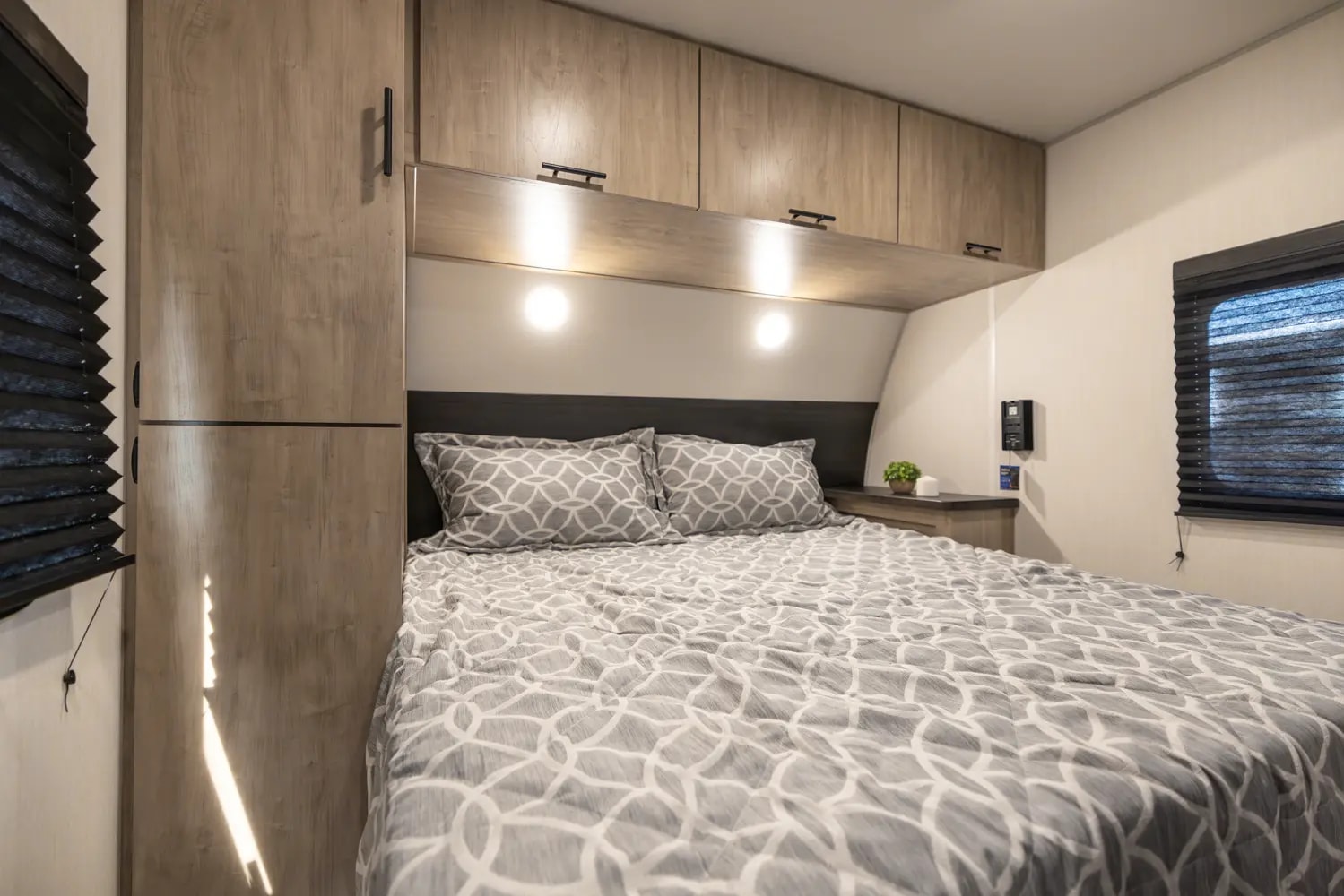 Inside a modern RV rental bedroom featuring a patterned bed, wood cabinets, reading lights, and a window with blinds.. Forest River Cherokee Grey Wolf 2025