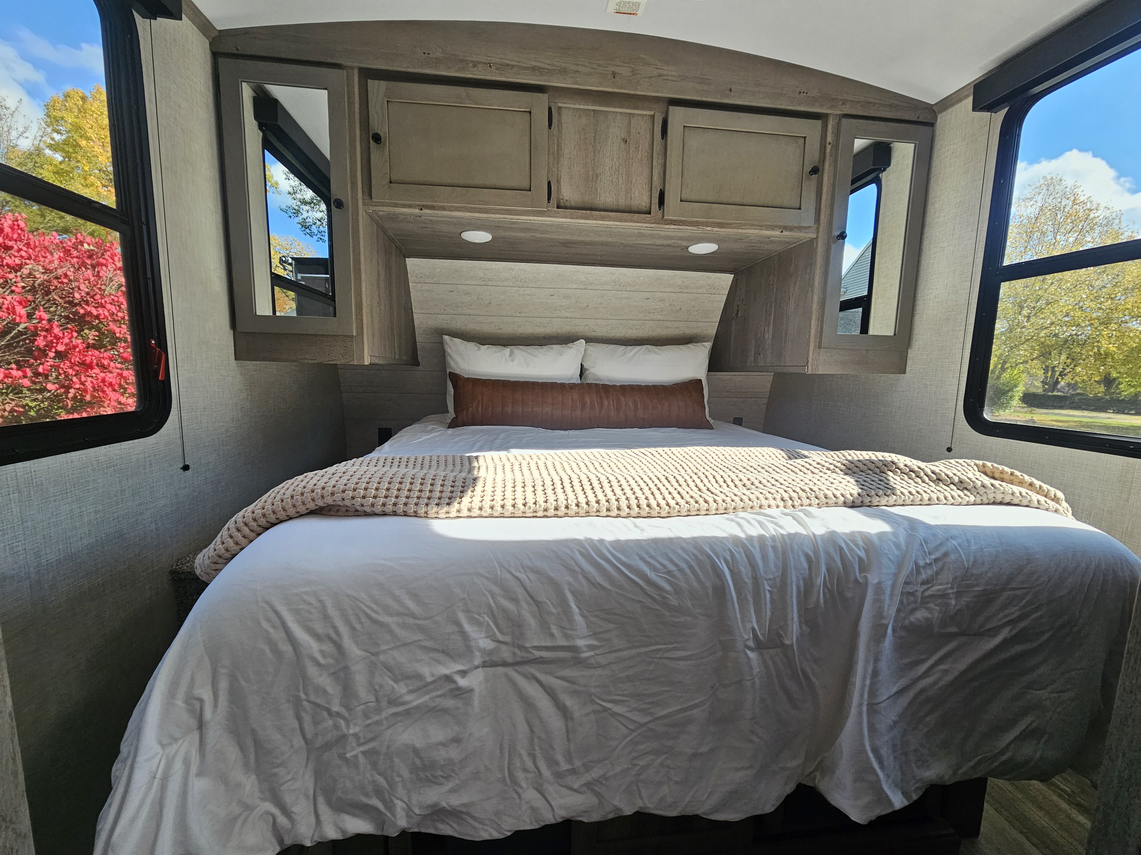 An inviting RV rental bedroom with a comfortable bed, overhead cabinets, and large windows showcasing vibrant autumn colors.. Keystone RV Outback Ultra-Lite 2021