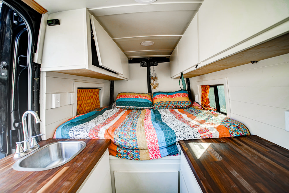 RV rental camper van interior featuring a cozy bed with colorful bedding, a sink, and wooden counters.. Mercedes-Benz Sprinter 2019