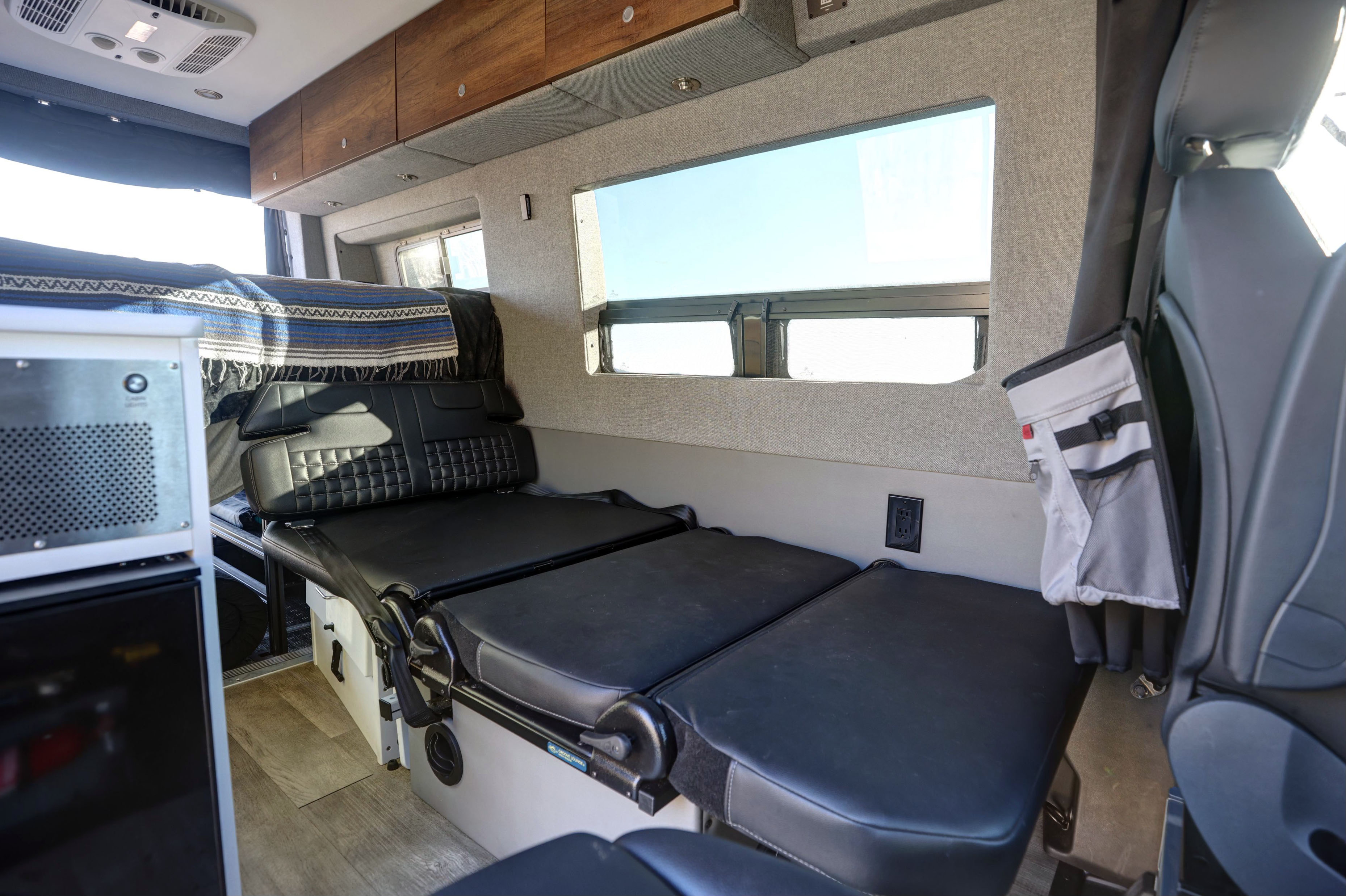 RV rental's versatile interior featuring convertible black leather seating/bed, wood overhead cabinets, and large windows.. Mercedes-Benz Sprinter 2021