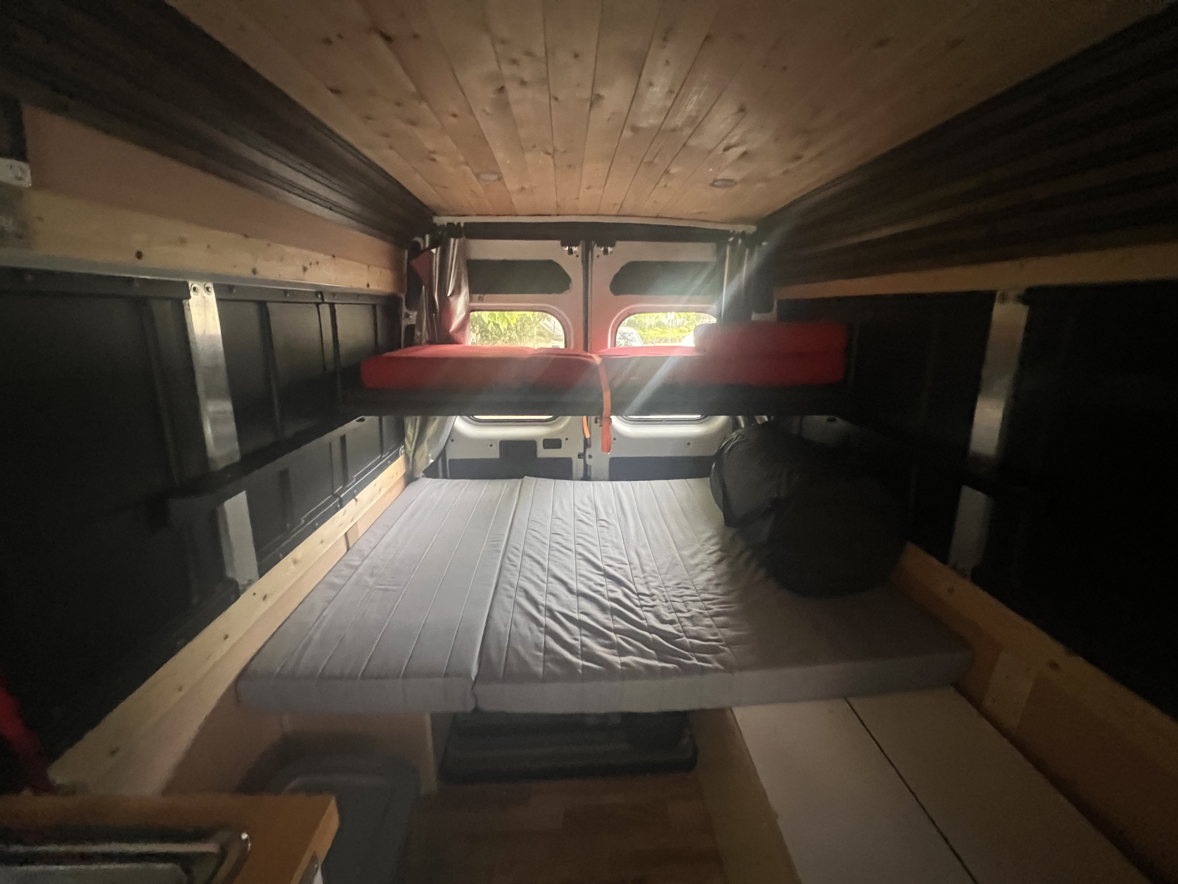 RV rental's inviting interior. Features a lower grey bed and an upper red bunk, wood ceiling, & natural light from rear windows.. Ram Promaster 2019