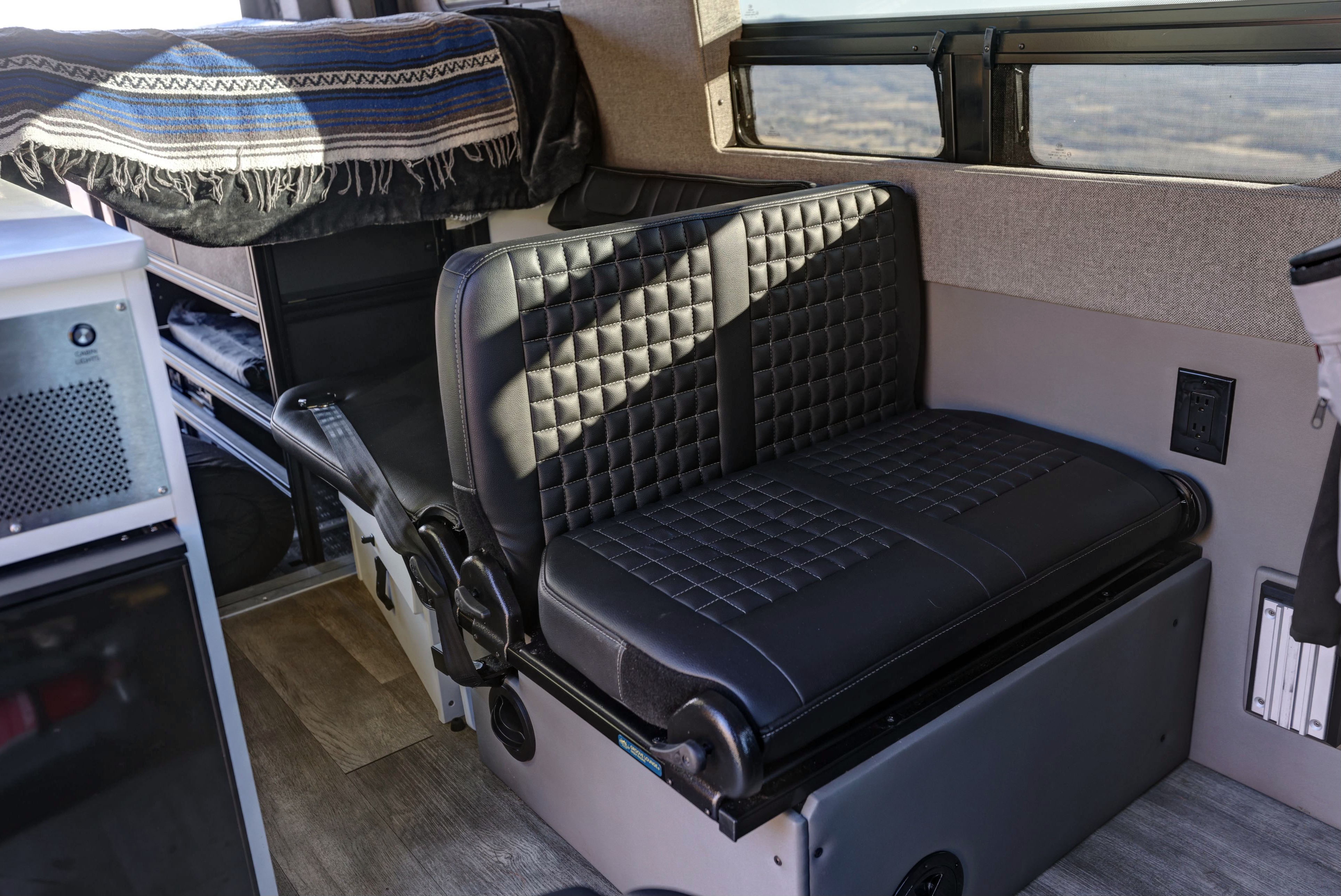 RV rental: Modern camper van interior featuring black quilted bench seats, a striped blanket, wood floor, and custom storage.. Mercedes-Benz Sprinter 2021