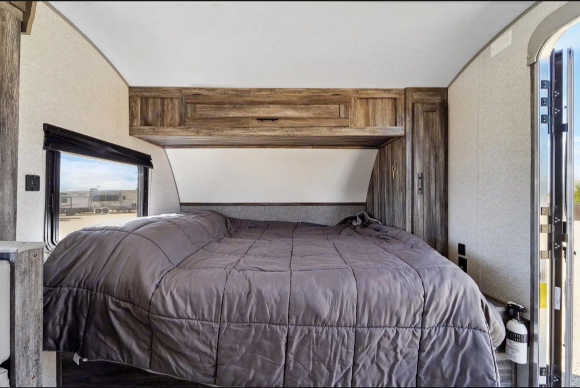 Comfortable RV rental bedroom featuring a large bed with a grey comforter, overhead wooden storage, window view, and an open door on the right.. Forest River Salem FSX 2021
