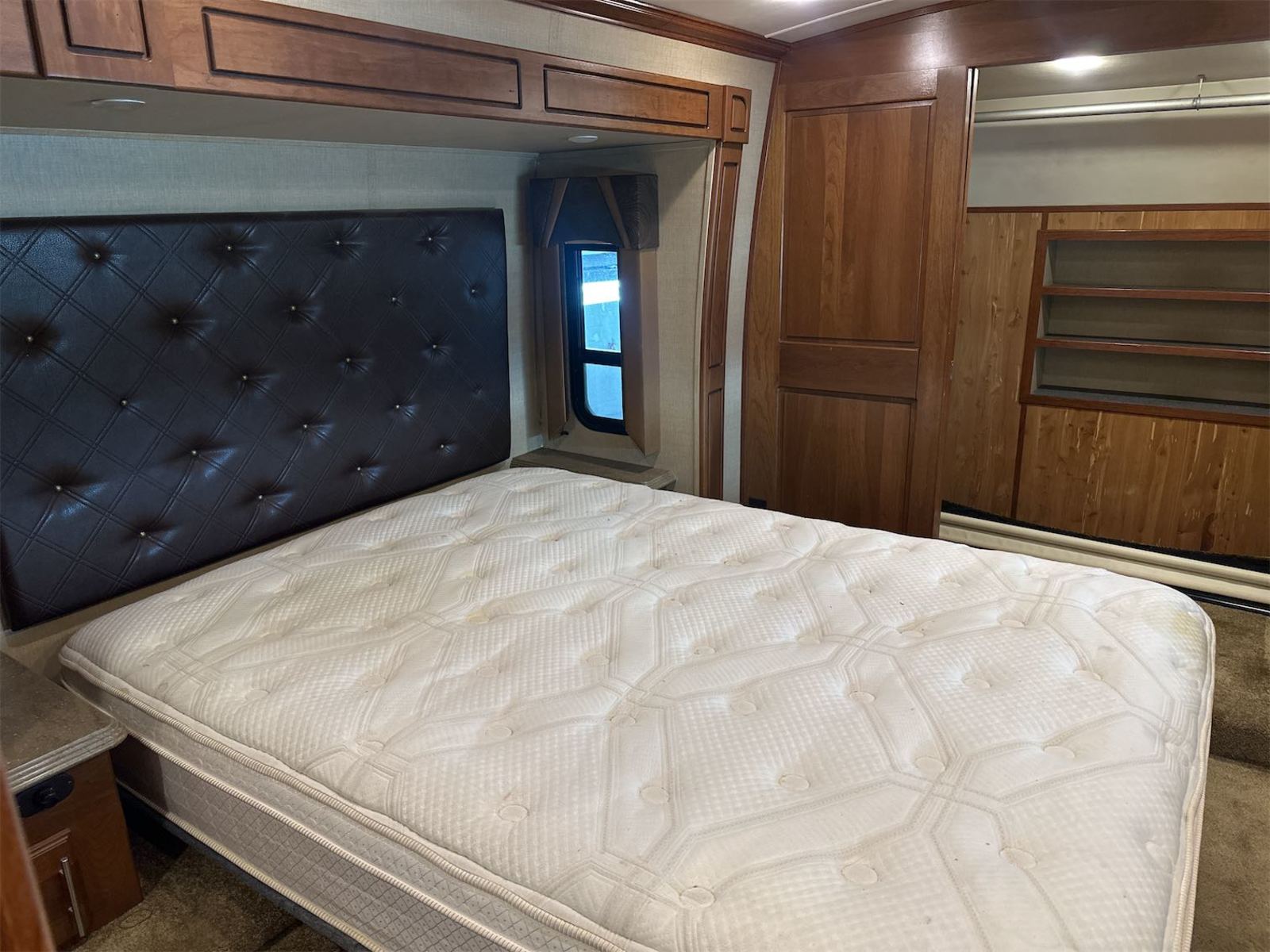 Spacious RV rental bedroom with a large mattress, dark tufted headboard, wooden storage, and window, offering a cozy sleep space.. Dynamax Trilogy 3850RL 2014