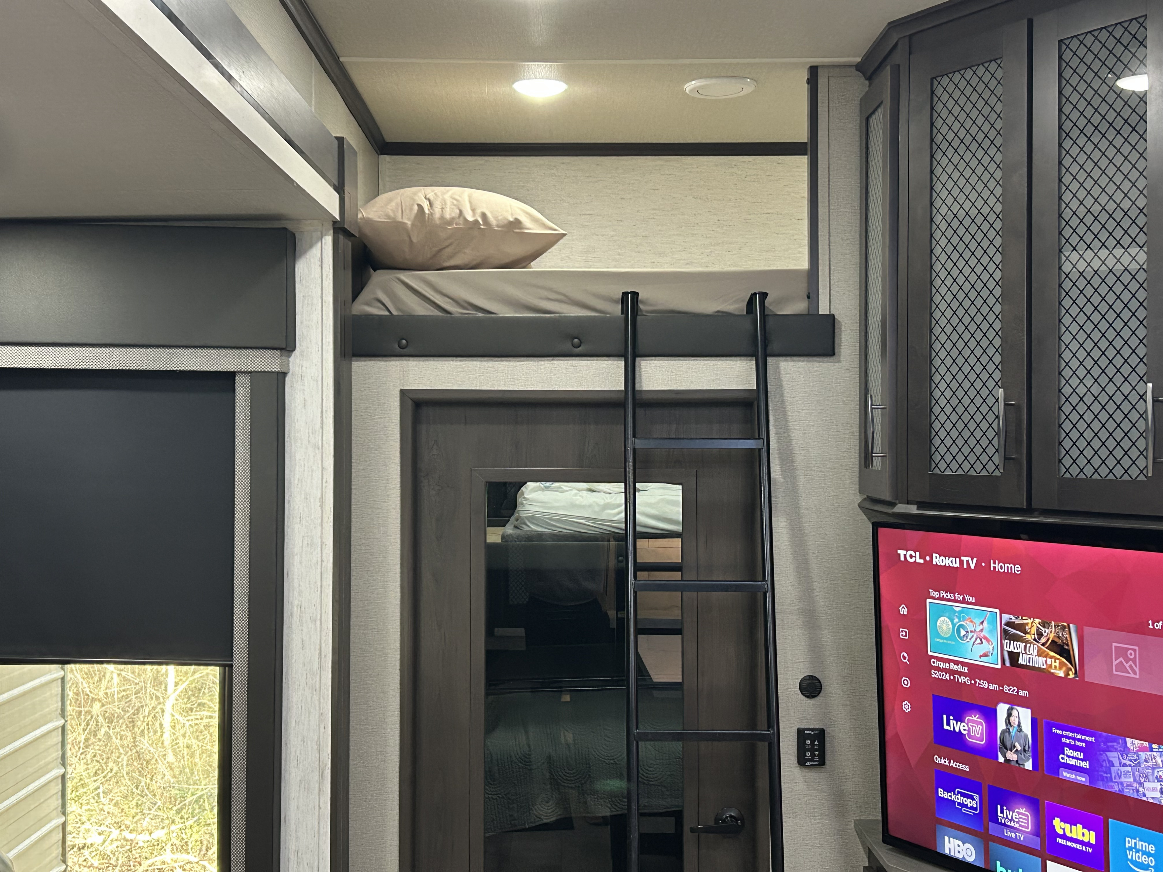 Inside an RV rental, a cozy bunk bed with a ladder, TV, and cabinets. A window with a shade shows trees outside.. Grand Design Momentum 2022