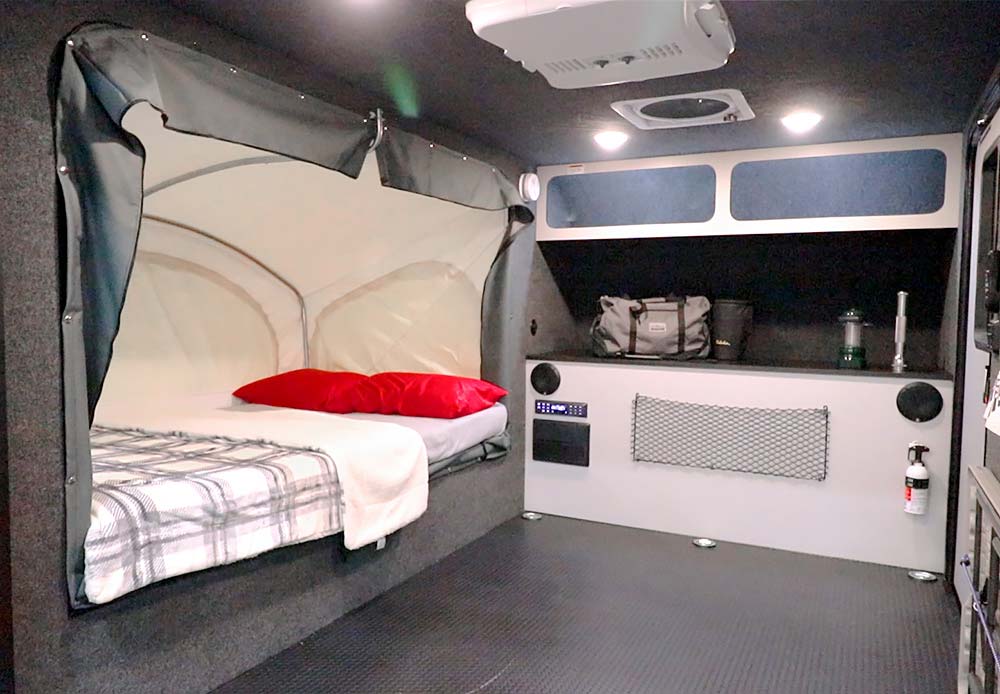 Compact RV rental interior featuring a cozy bed with red pillows, plaid blanket, storage shelves, and utility panel. Adventure awaits!. IntechRV Flyer Explore 2021