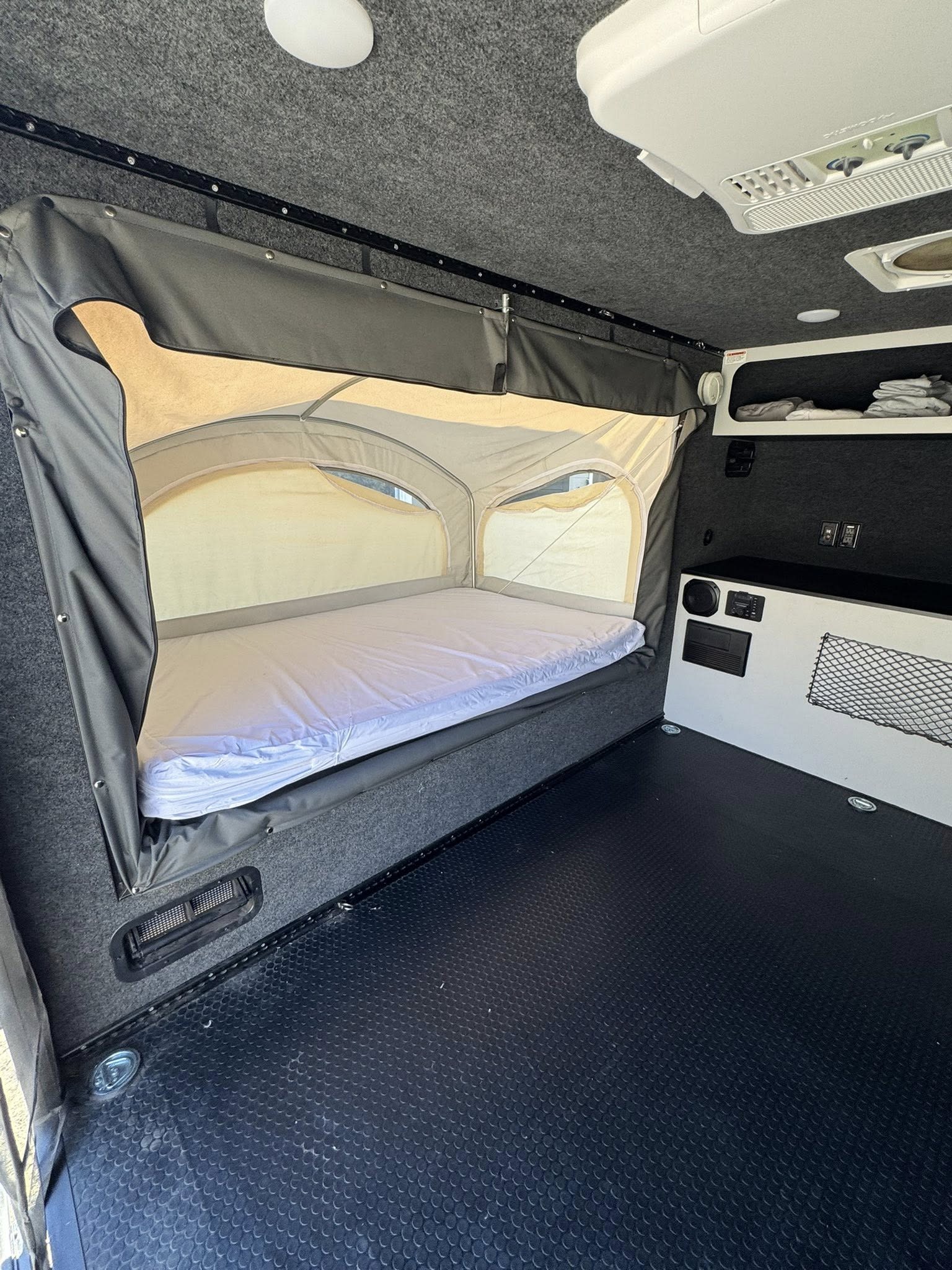 RV rental interior: a deployable bed with white mattress, textured black flooring, and an overhead AC unit for comfort.. IntechRV Flyer Explore 2021