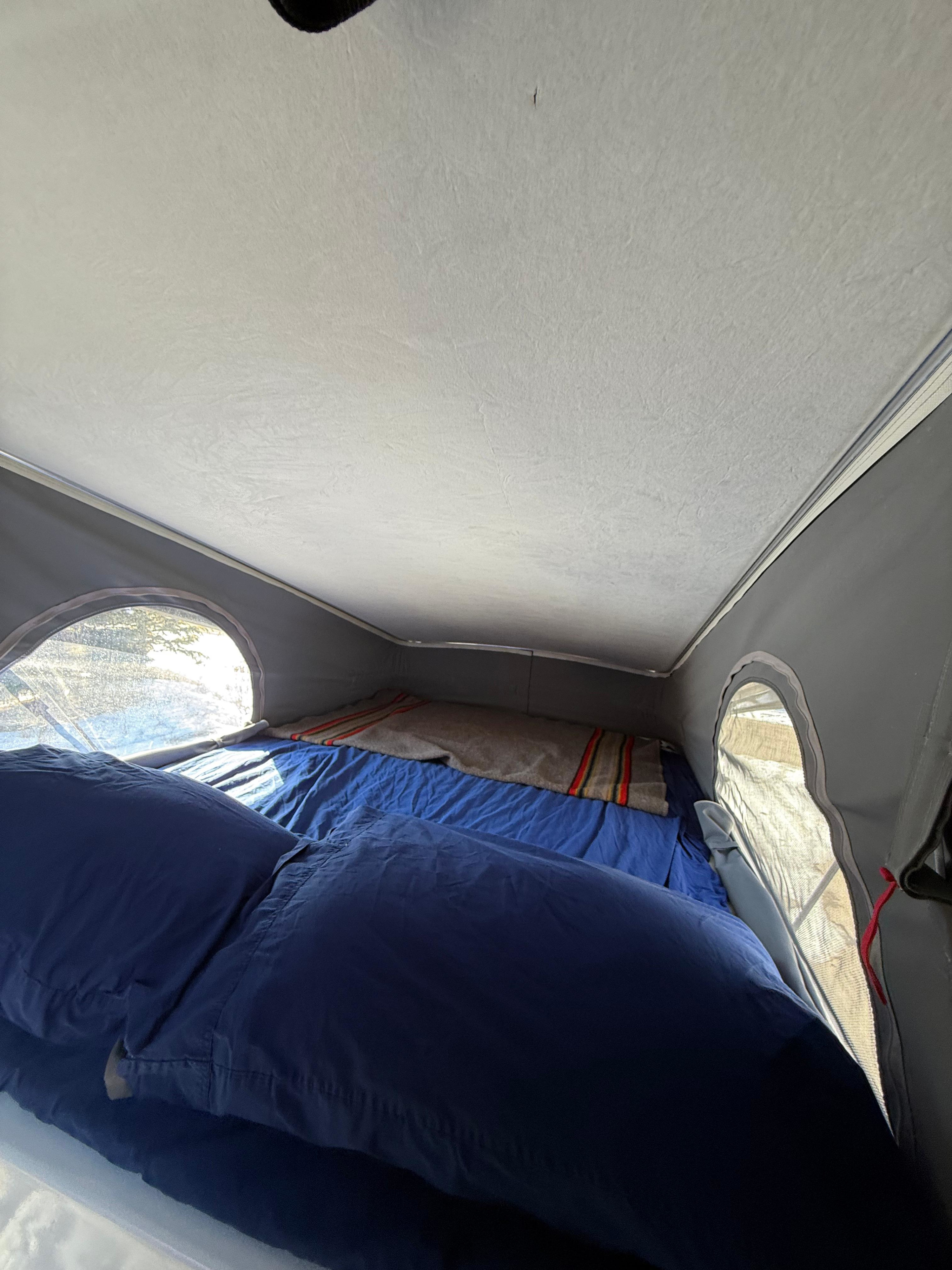 RV rental pop-top interior: a cozy bed with blue bedding and a striped blanket, illuminated by side windows.. Winnebago Solis 2021