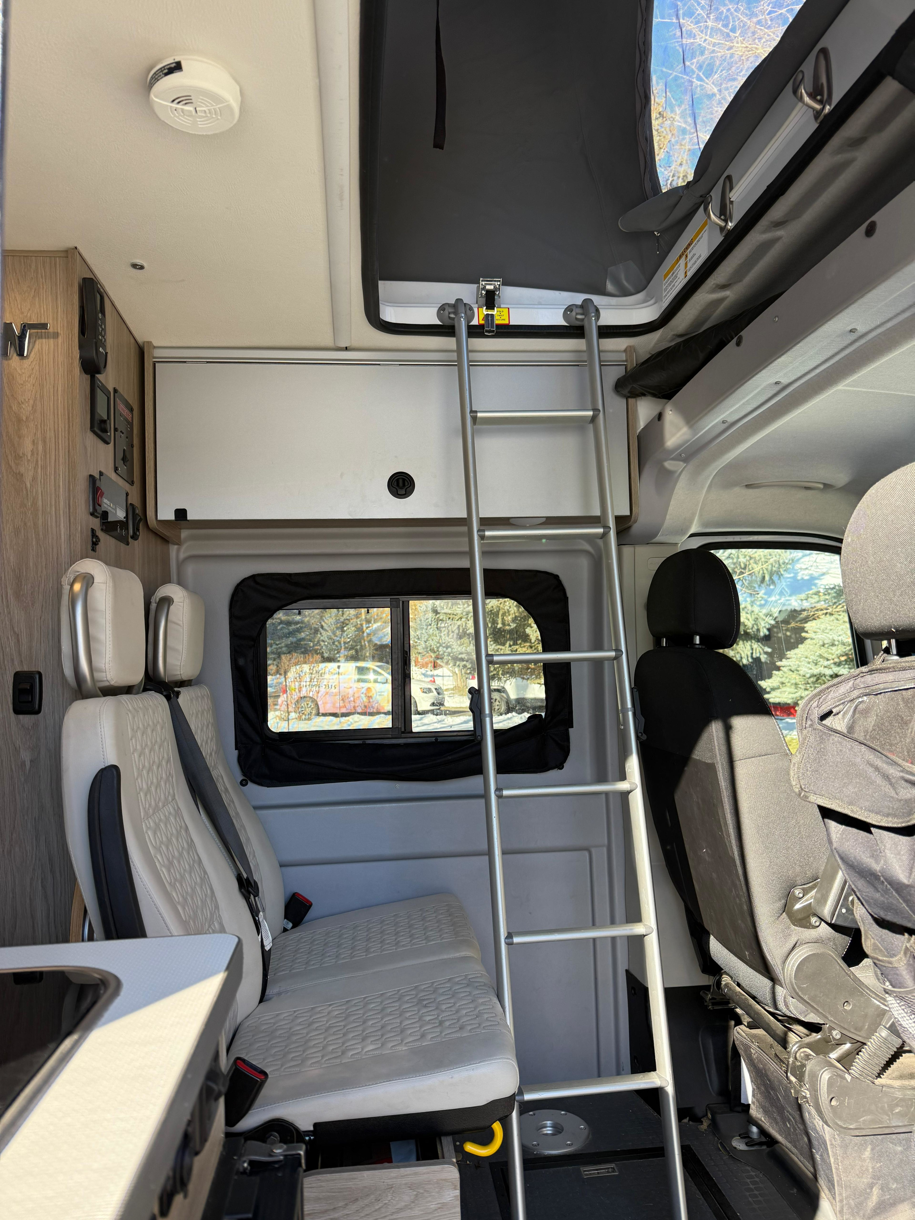 Interior of an RV rental: ladder to a pop-top bunk, passenger seats, wall controls, and a winter window view.. Winnebago Solis 2021