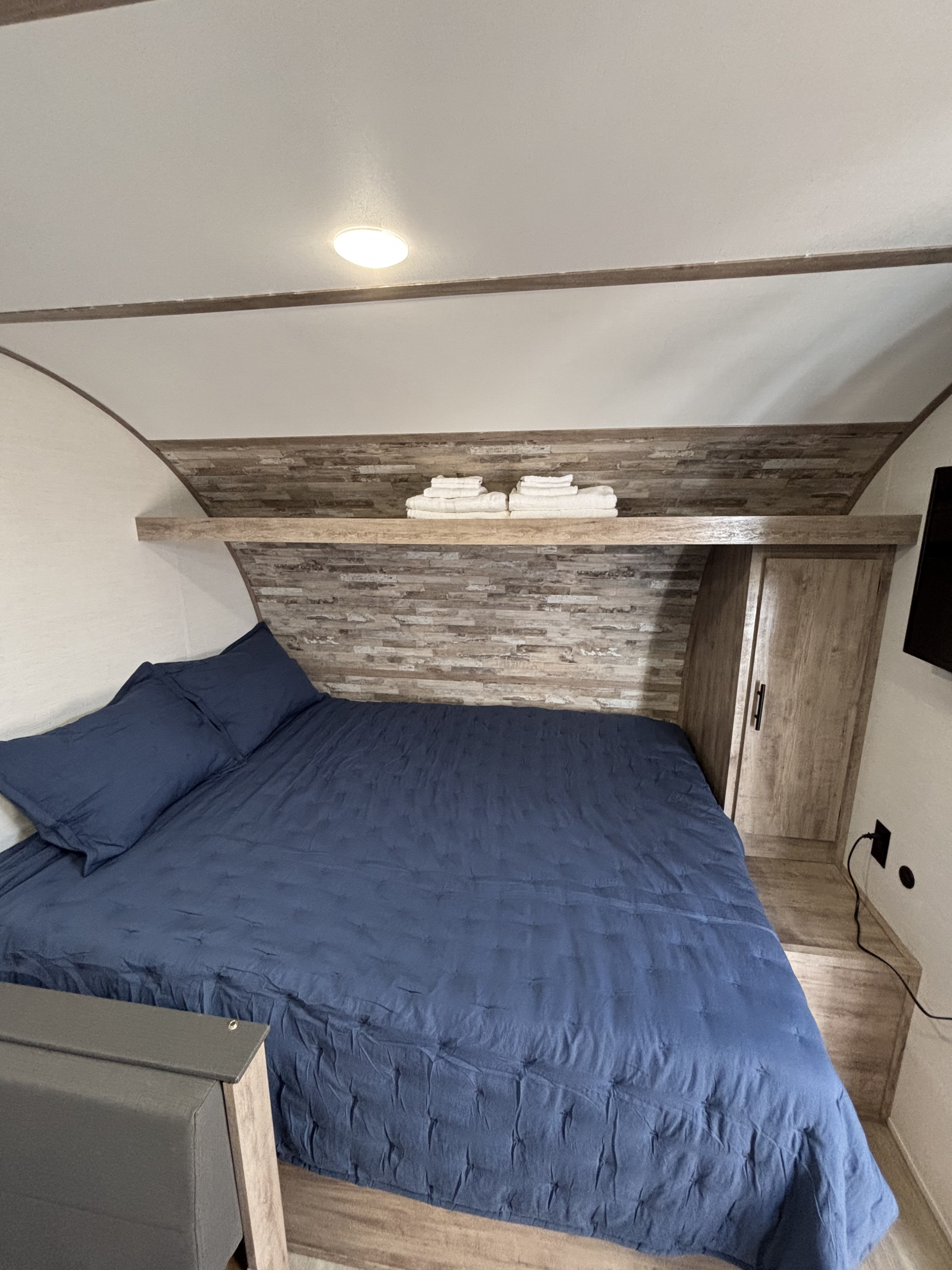 Cozy rv rental bedroom featuring a blue bed, textured wood accent wall, shelf with towels, and built-in storage.. Gulf Stream Enlighten Super Lite 2025
