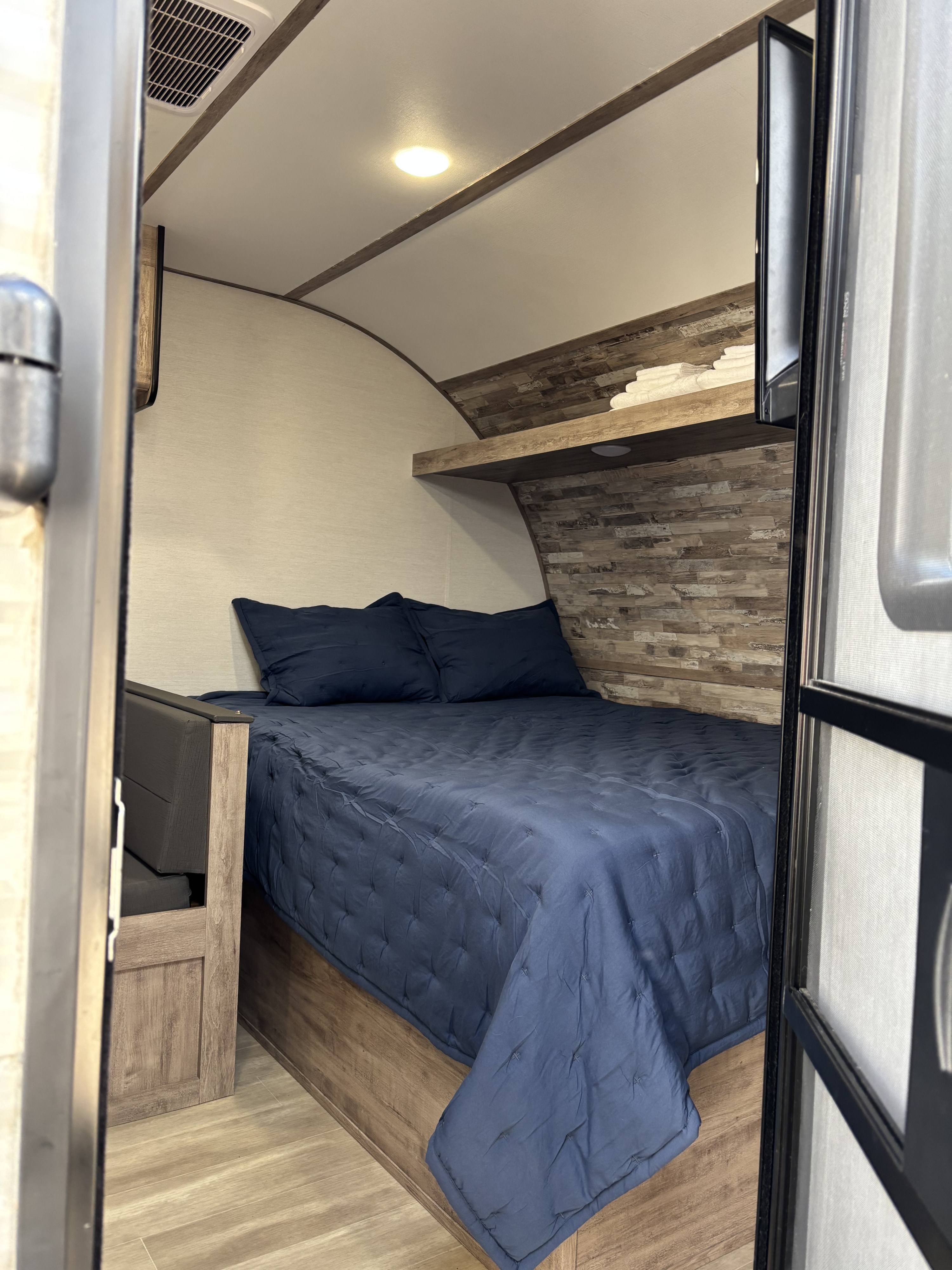 Cozy RV rental bedroom with a blue quilted bed, wood-paneled wall, overhead light, and AC vent, viewed from the doorway.. Gulf Stream Enlighten Super Lite 2025