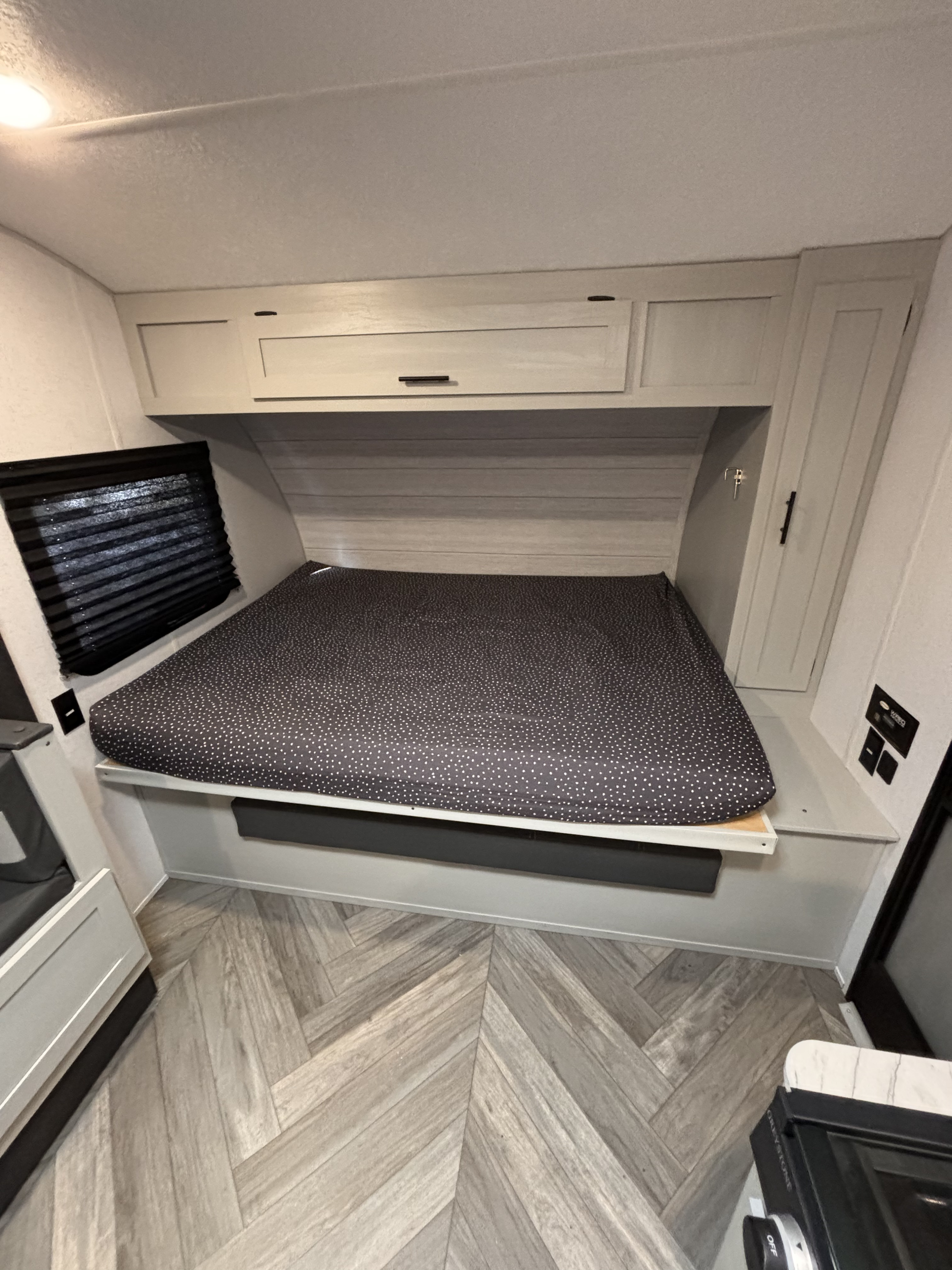 Cozy RV rental bedroom with a spotted mattress, overhead cabinets, a window, and light wood-look herringbone flooring.. Forest River Wildwood FSX 2022