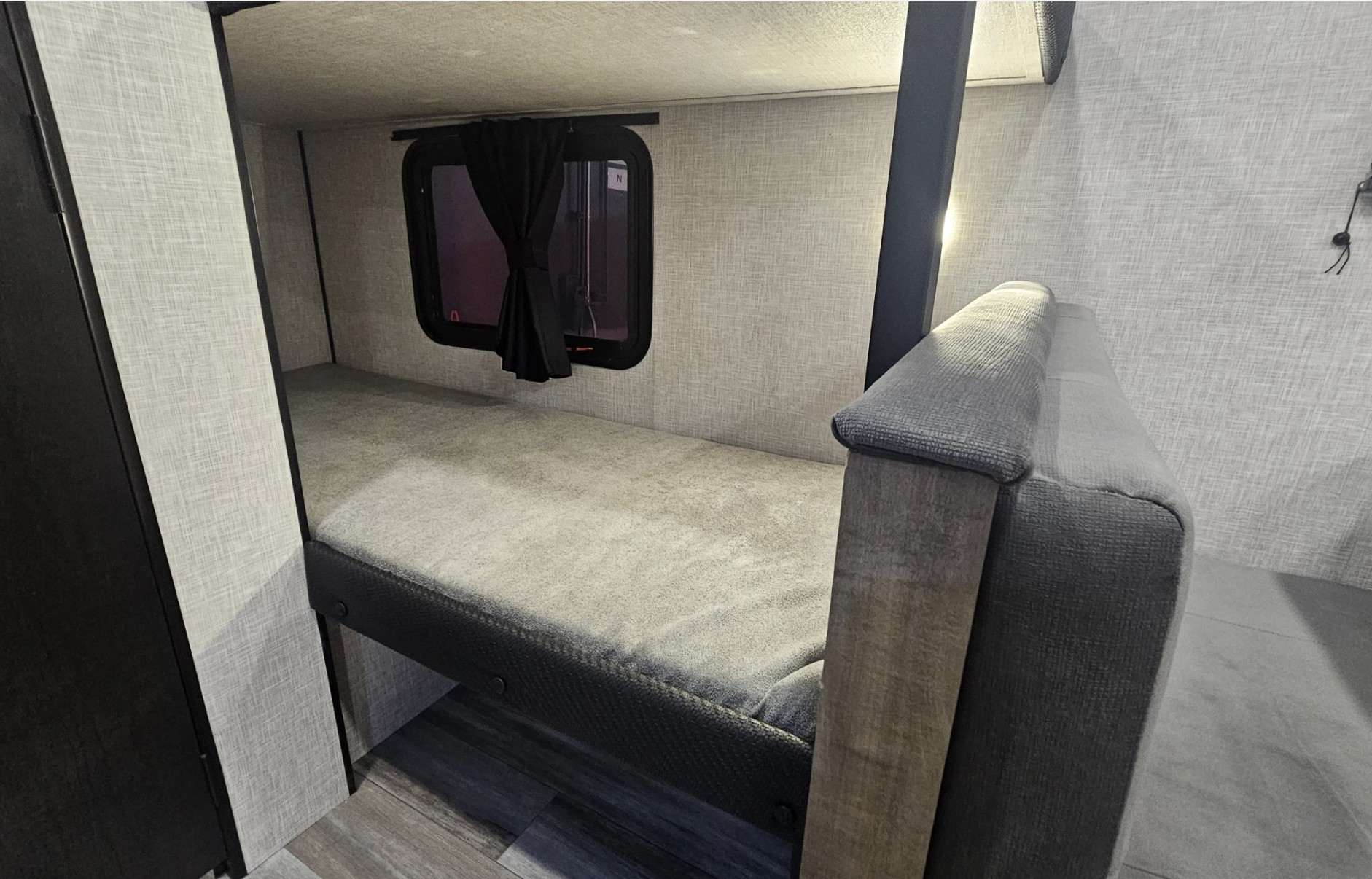 RV rental bunk bed area: gray mattress, small window with black curtains, textured walls, and part of a comfy sofa.. Jayco JayFlight 2026