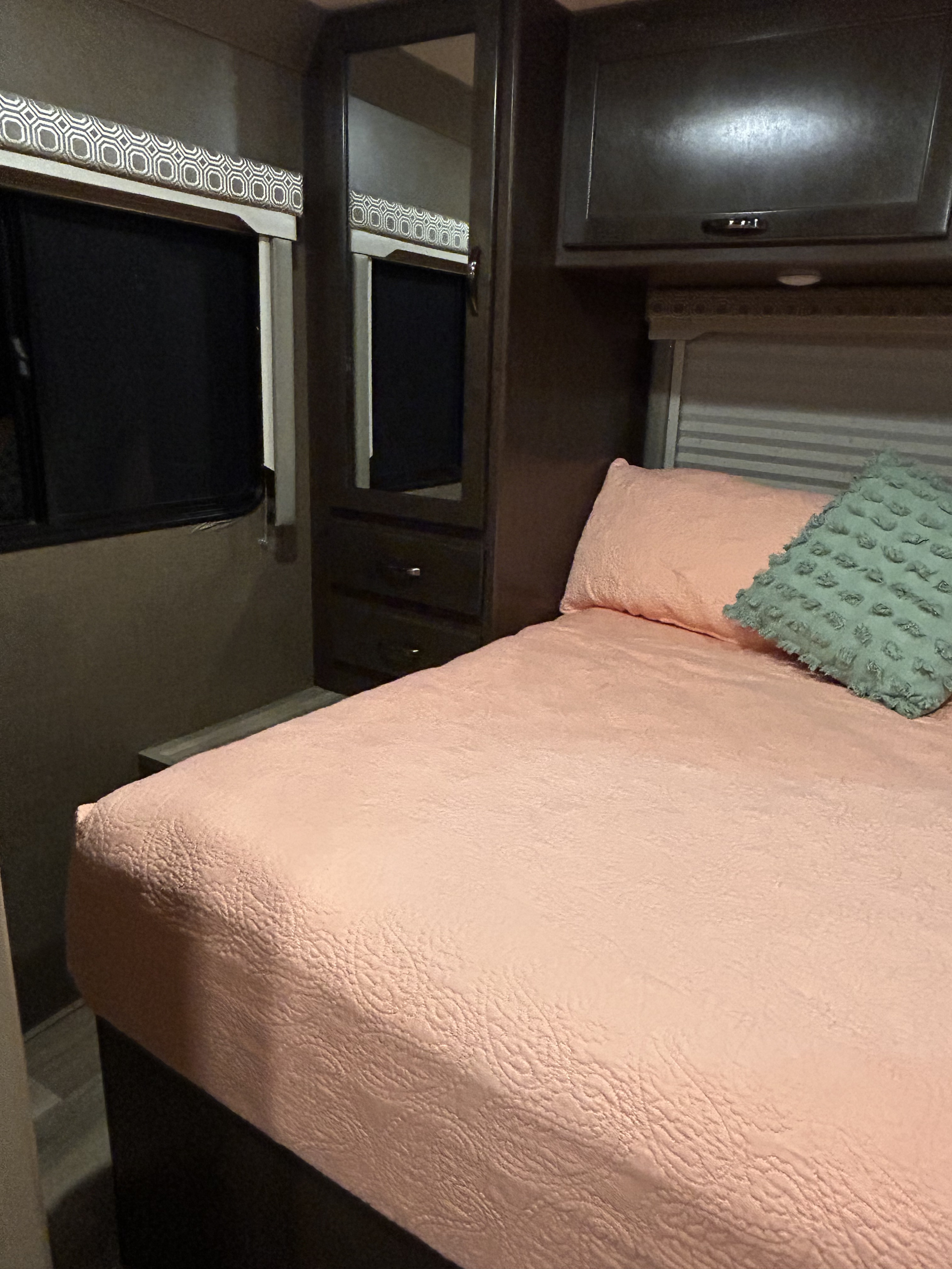 Cozy RV rental bedroom with peach bed, accent pillow, dark cabinets, mirrored wardrobe, and window with patterned valance.. Thor Four Winds 2020