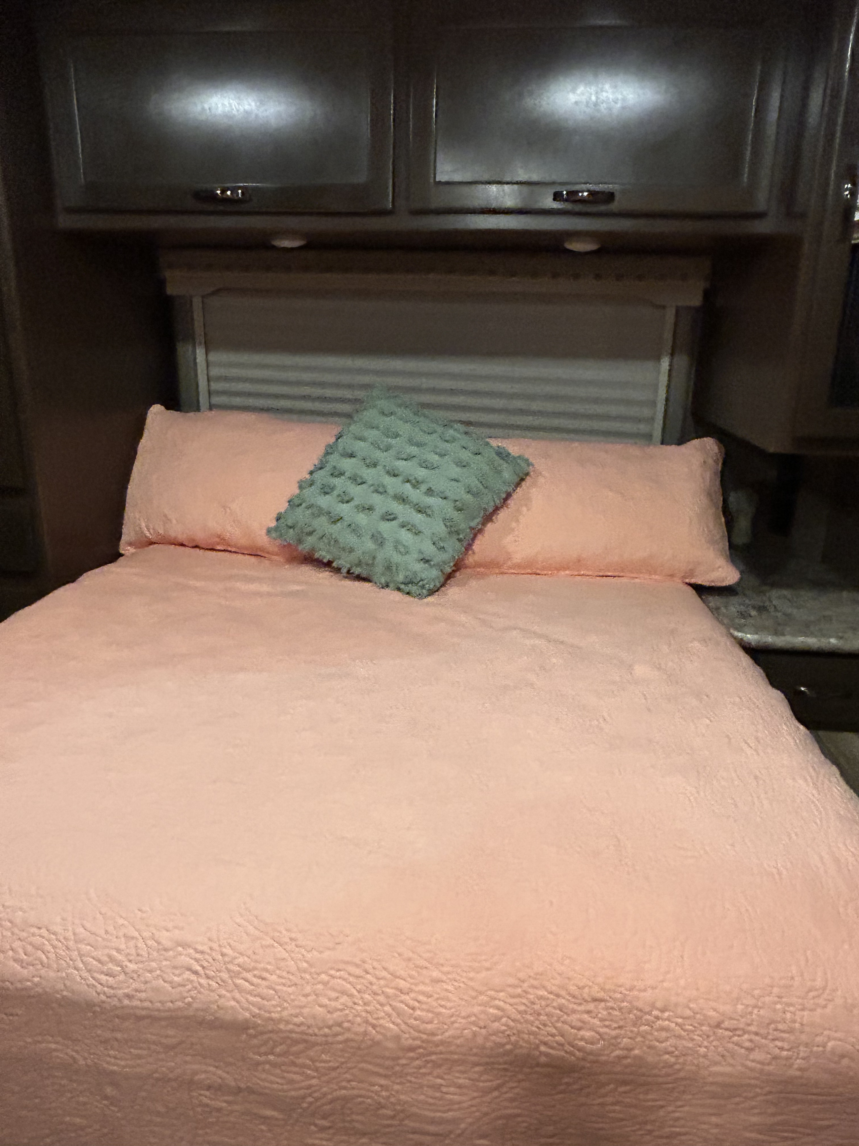 An RV rental bedroom with a comfortable bed, pink comforter, pillows, and a textured green accent pillow under dark cabinets.. Thor Four Winds 2020