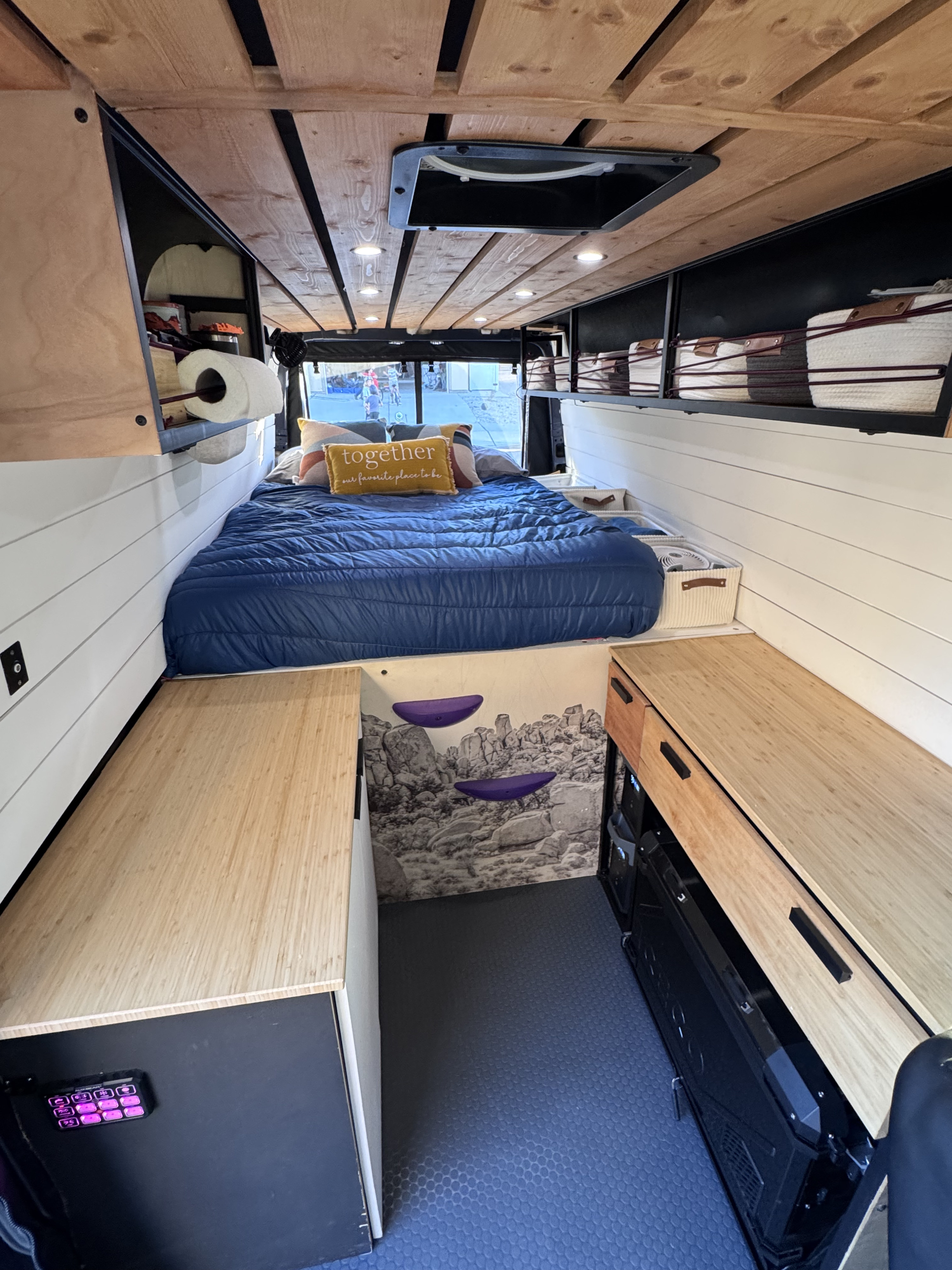 Cozy rv rental interior featuring a bed, wood-slat ceiling, white shiplap walls, storage, and a unique rock climbing wall graphic.. Mercedes-Benz Sprinter 2021