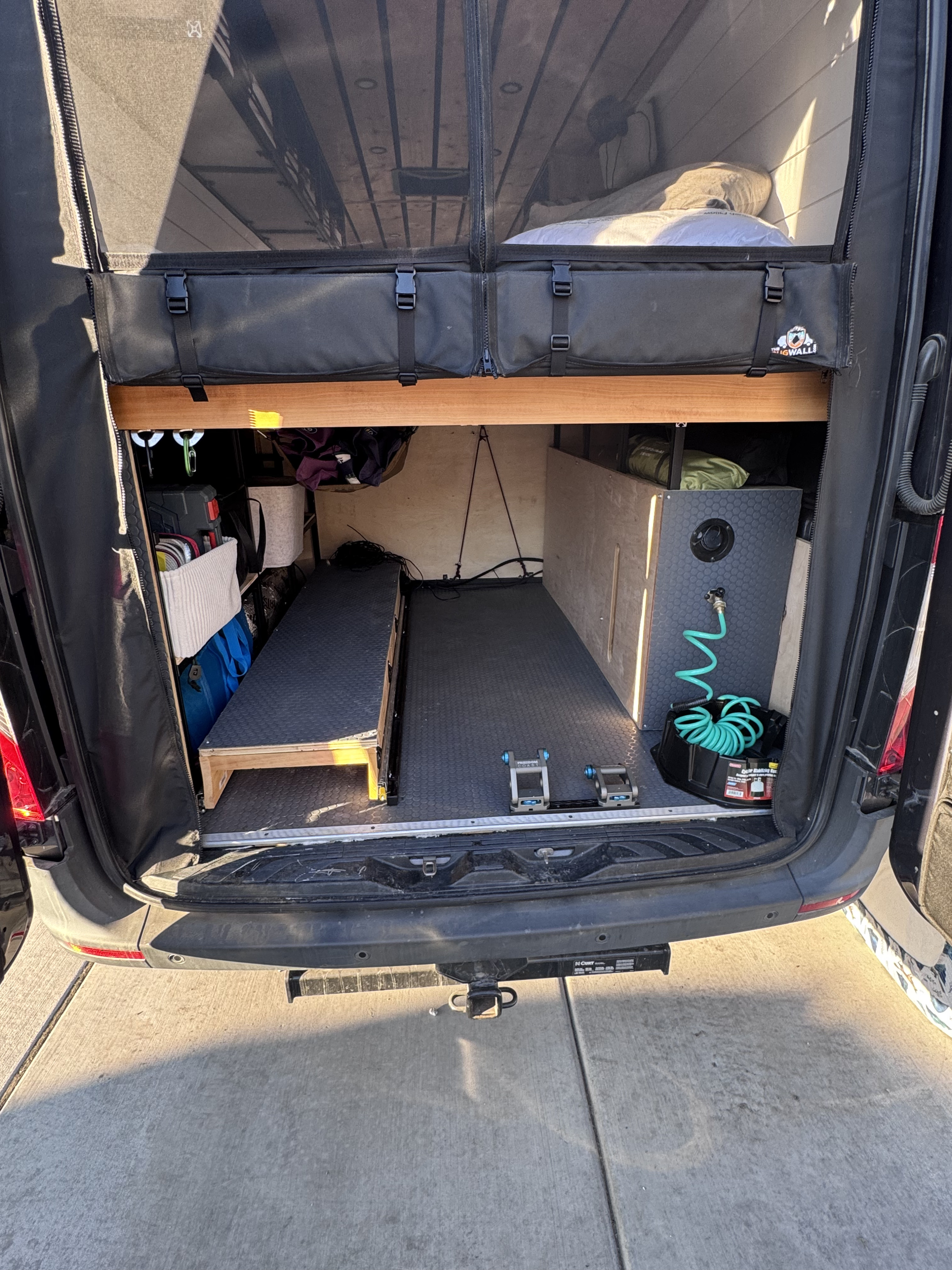 Rear view of an RV rental camper van's organized interior: storage, bed platform, water spigot, and bug screen.. Mercedes-Benz Sprinter 2021