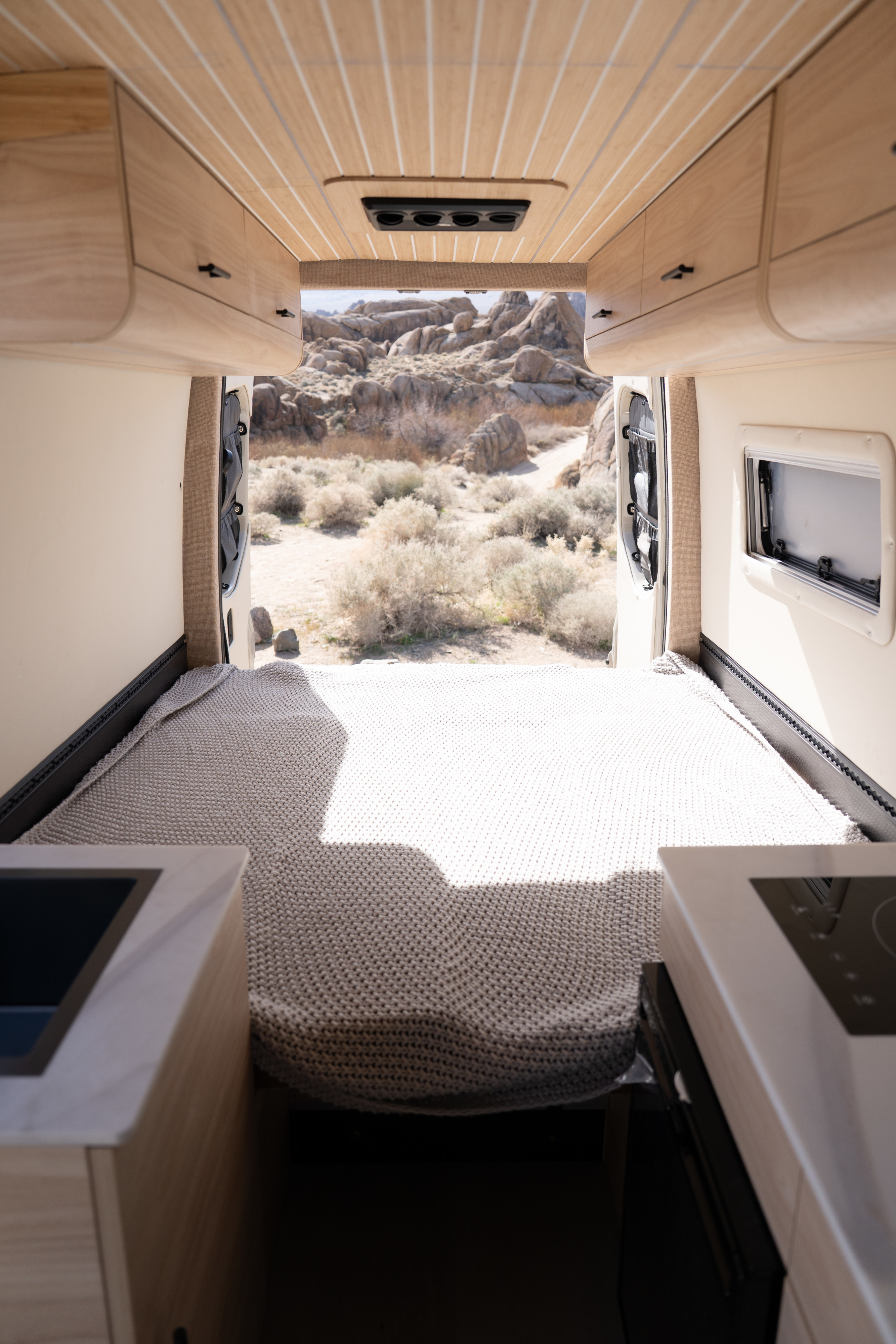RV rental: Cozy bed, wood interior, and open rear doors reveal a stunning desert landscape. A kitchenette is visible foreground.. Sandy Vans Mercedes Sprinter 2025