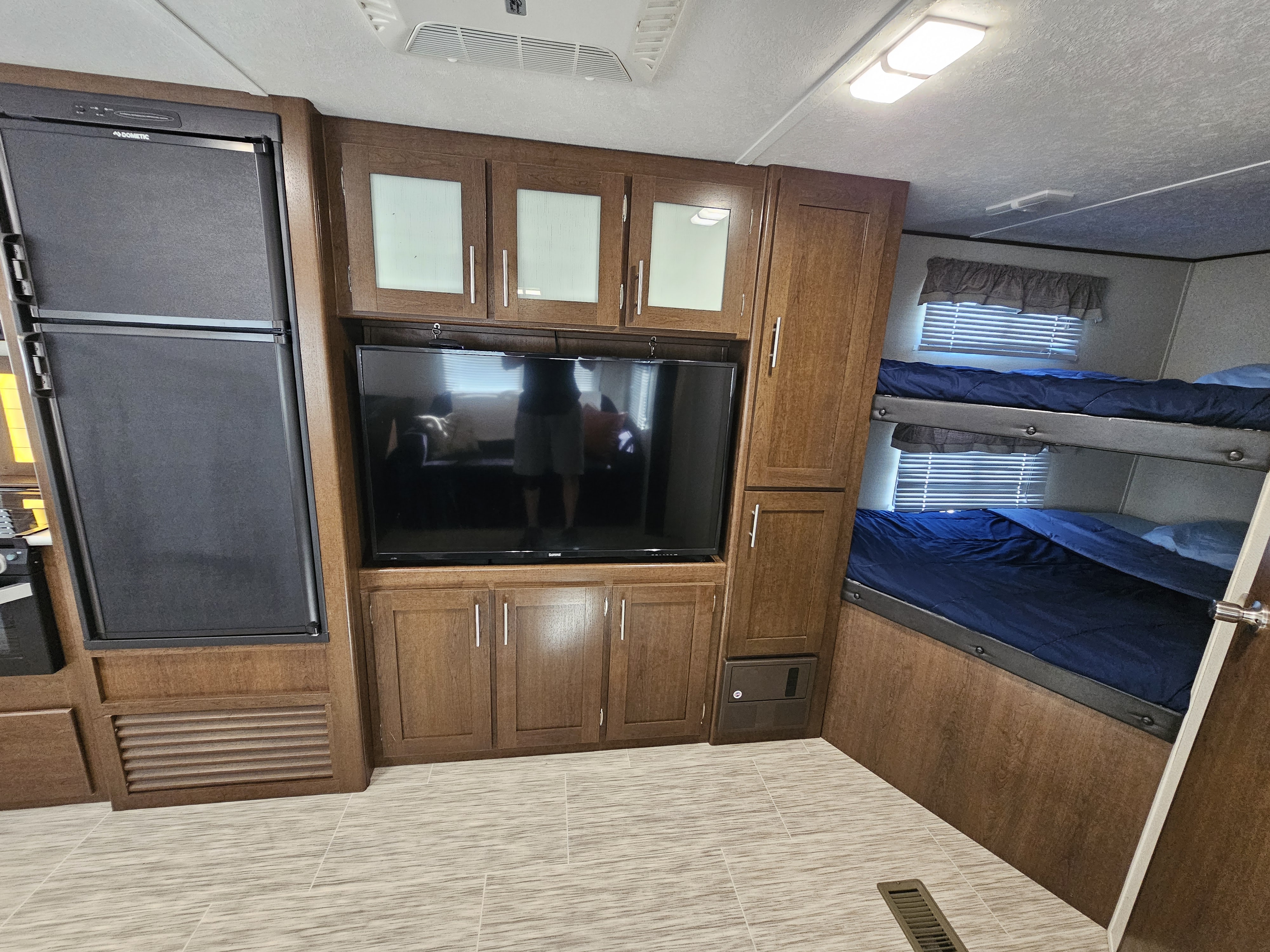 An RV rental's cozy interior, showing a black Dometic fridge, TV, dark wood cabinets, and two bunk beds with blue bedding.. Avenger ATI 2019