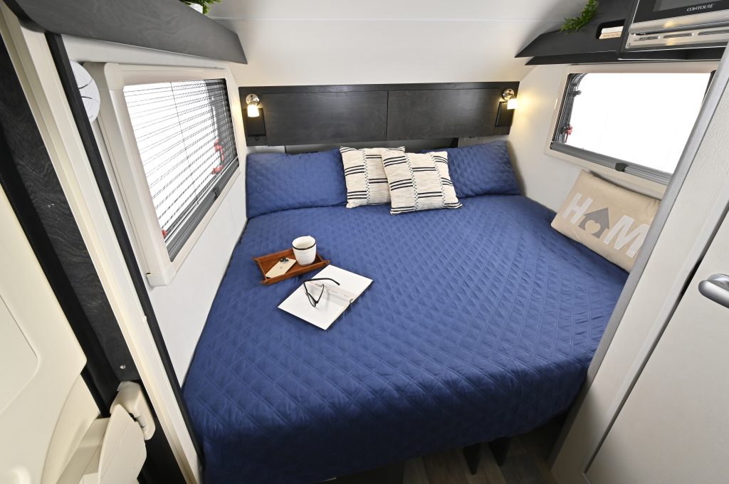 Cozy RV rental bedroom with a large bed, blue quilted bedding, two windows, and a reading setup for comfortable journeys.. nuCamp T@B 2023