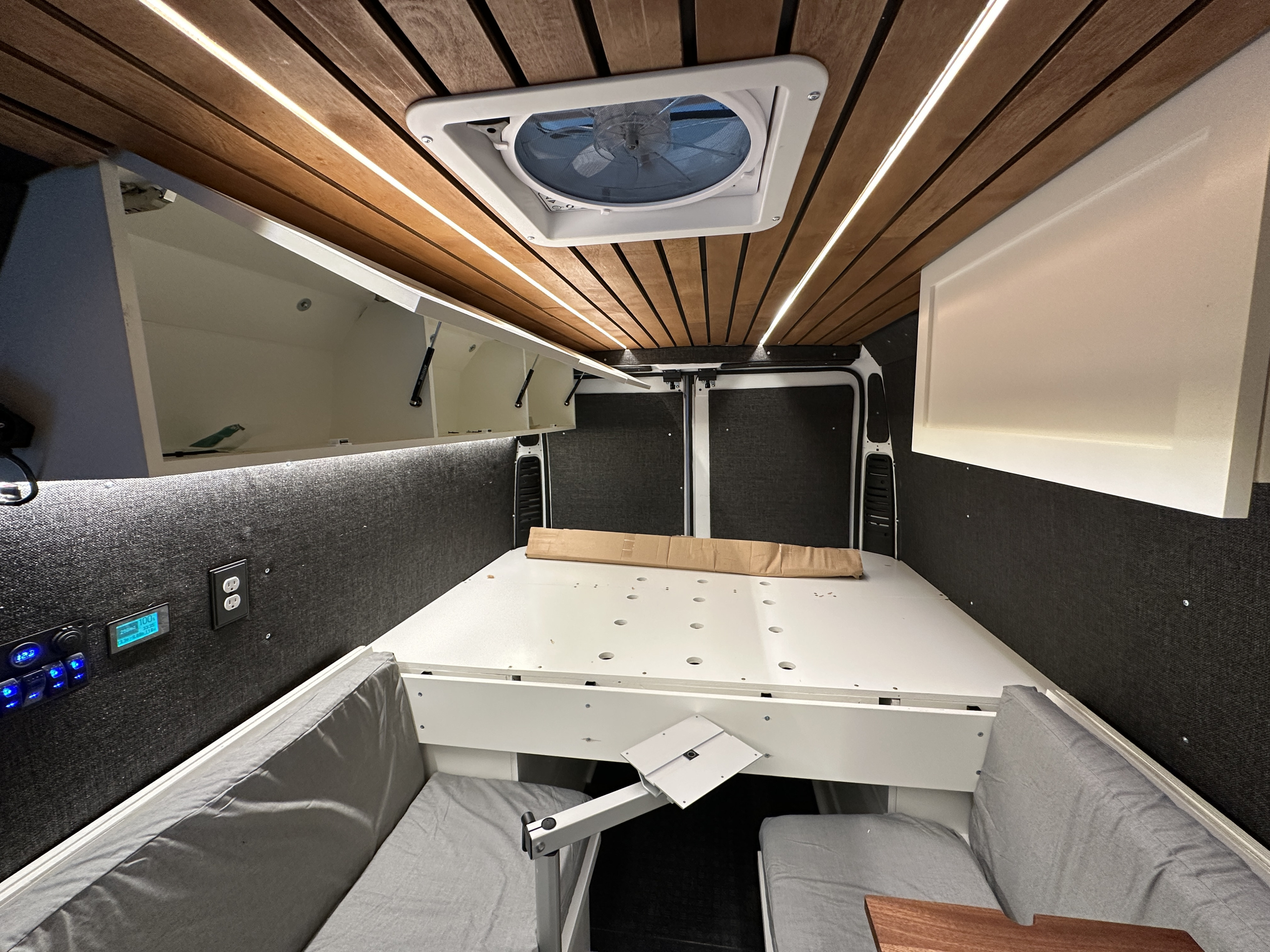 Inside a sleek rv rental van: wood ceiling with LED strips, fan, bed, control panel, & comfy benches.. Dodge B Van 2023