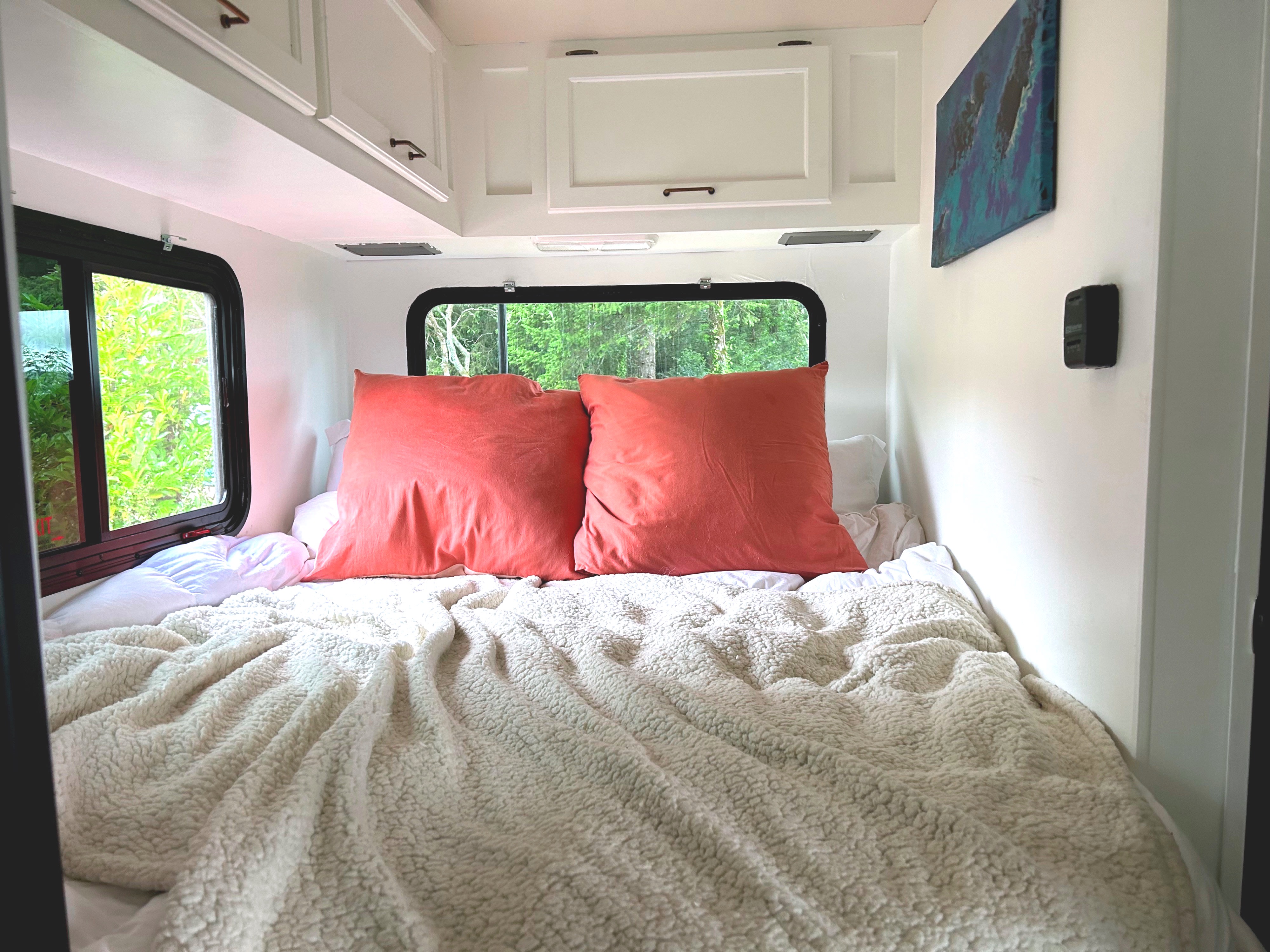 Cozy rv rental bedroom featuring a bed with coral pillows, a fluffy white blanket, white overhead cabinets, and green window views.. Thor Chateau 2016