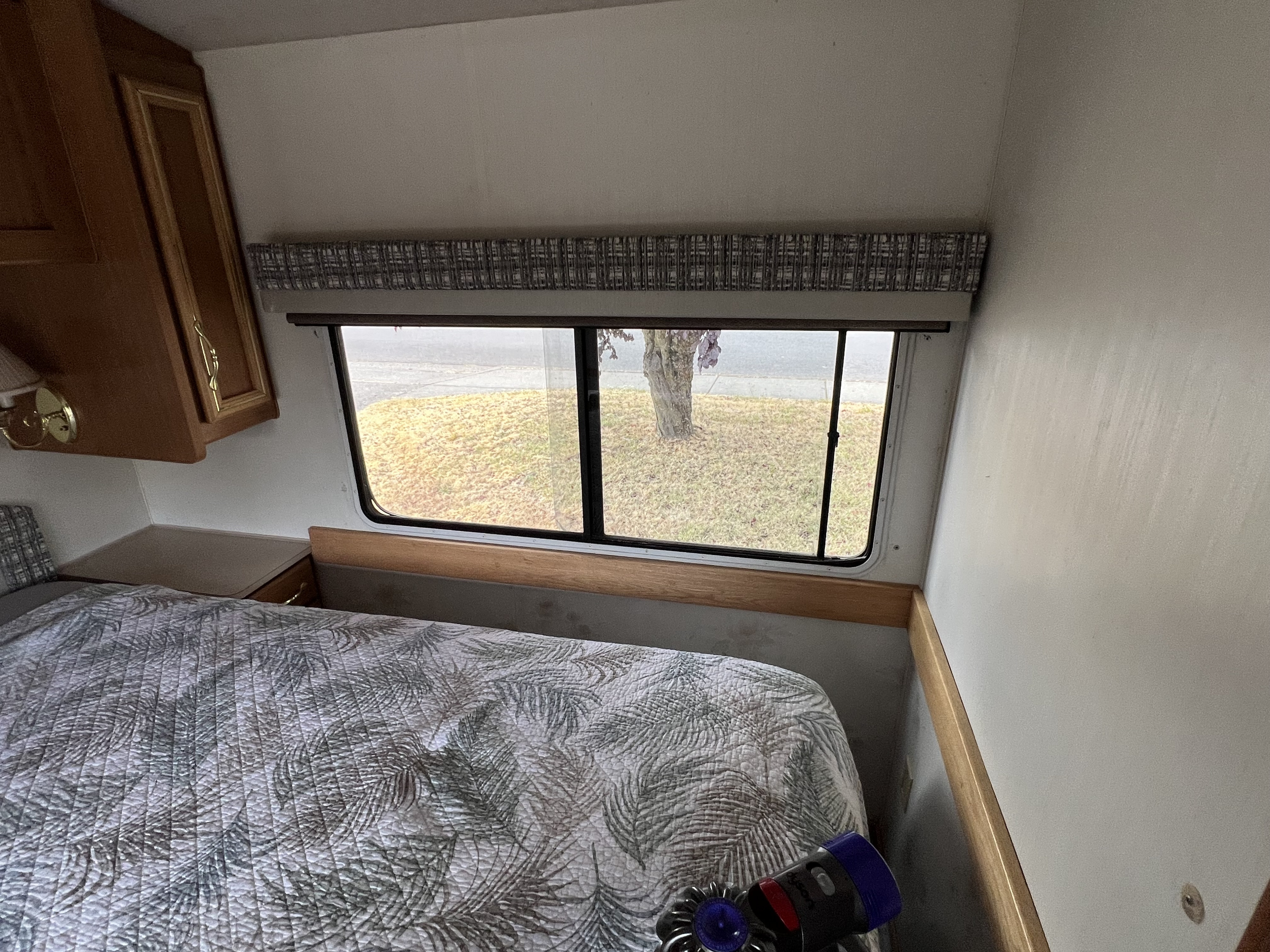 RV rental bedroom interior featuring a patterned bed, window view of a tree, and noticeable wall stains near the window.. Fleetwood Tioga Sl 1998