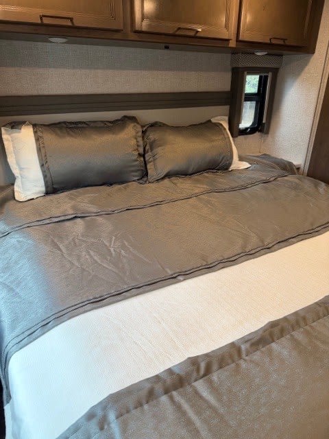 Comfortable RV rental bedroom featuring a neatly made bed with grey and white linens, overhead cabinets, and window.. Thor Hurricane 2022