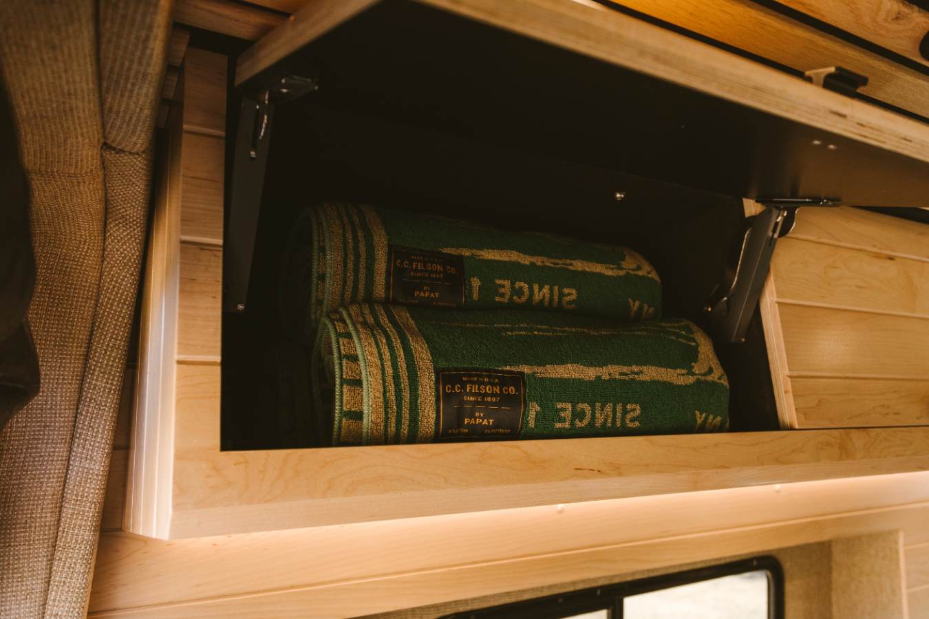 Organized overhead cabinet in an RV rental, featuring two green C.C. Filson blankets and warm wooden finishes.. Mercedes-Benz Sprinter 2025