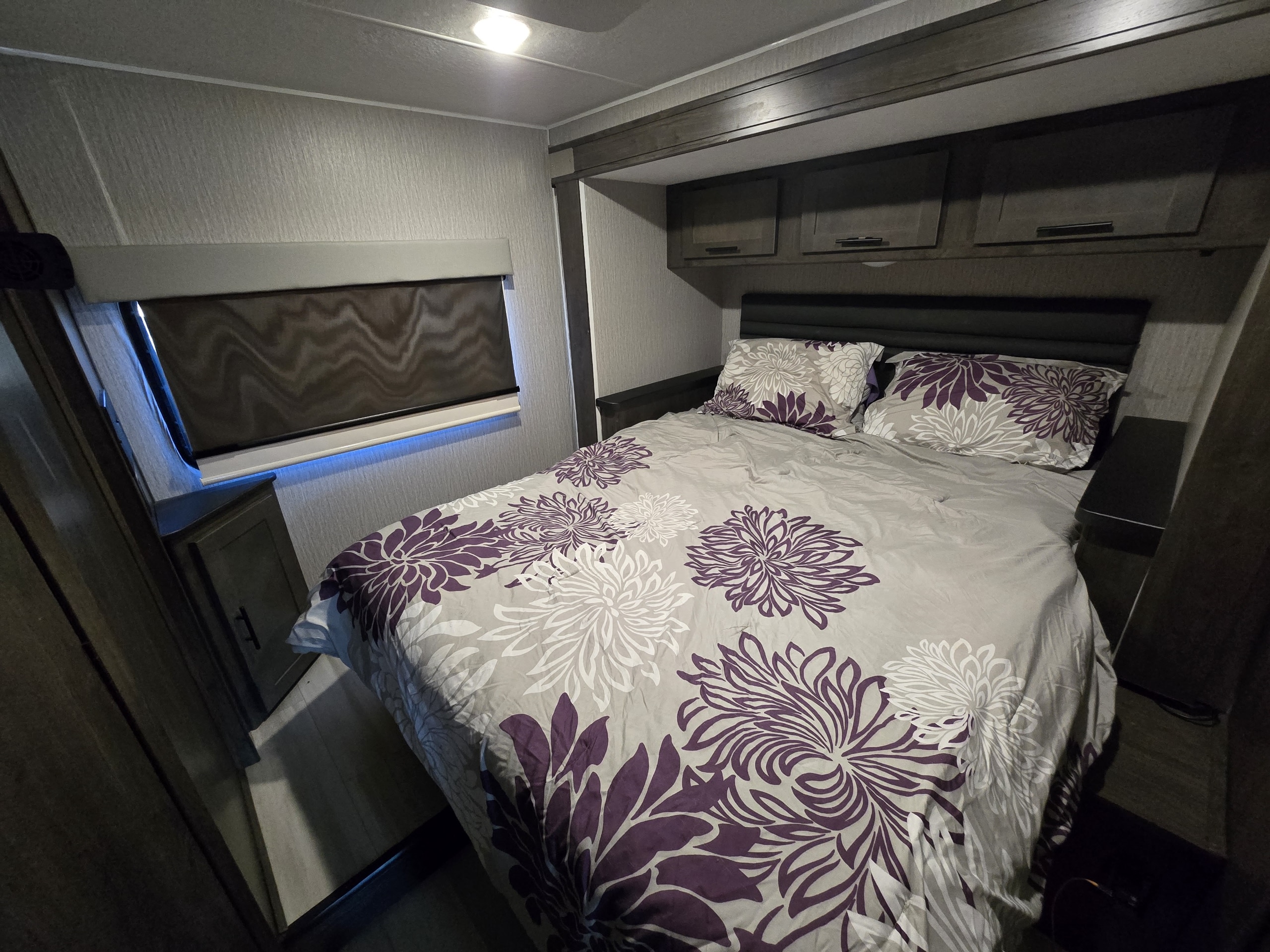 RV rental bedroom, offering a cozy bed with purple floral bedding, convenient overhead cabinets, and ambient window lighting.. Forest River Coachmen Leprechaun Premier 2022