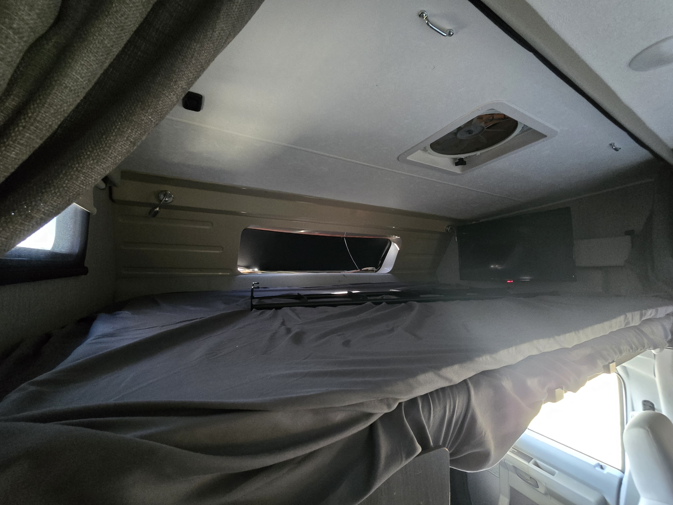 Inside an rv rental, showing the cozy overcab bunk bed with grey sheets, a TV, ceiling vent, and window.. Forest River Coachmen Leprechaun Premier 2022