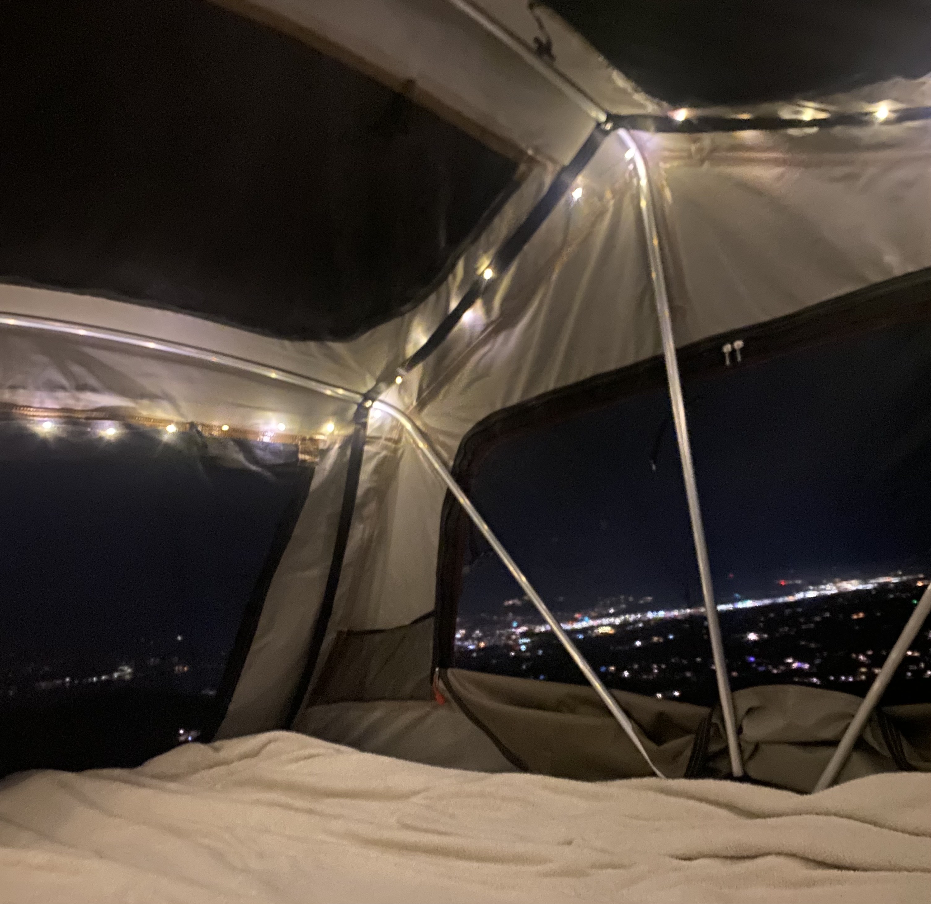 Inside a cozy rooftop tent at night, adorned with fairy lights, overlooking a twinkling city, perfect for an RV rental trip.. Toyota Rav4 2018