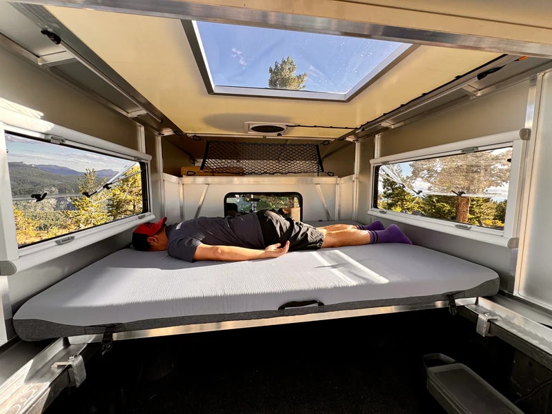 Scenic RV rental adventure: A man naps comfortably in a camper bed, enjoying mountain vistas from large windows and a skylight.. Chevrolet Silverado 1500 Trail Boss 2020